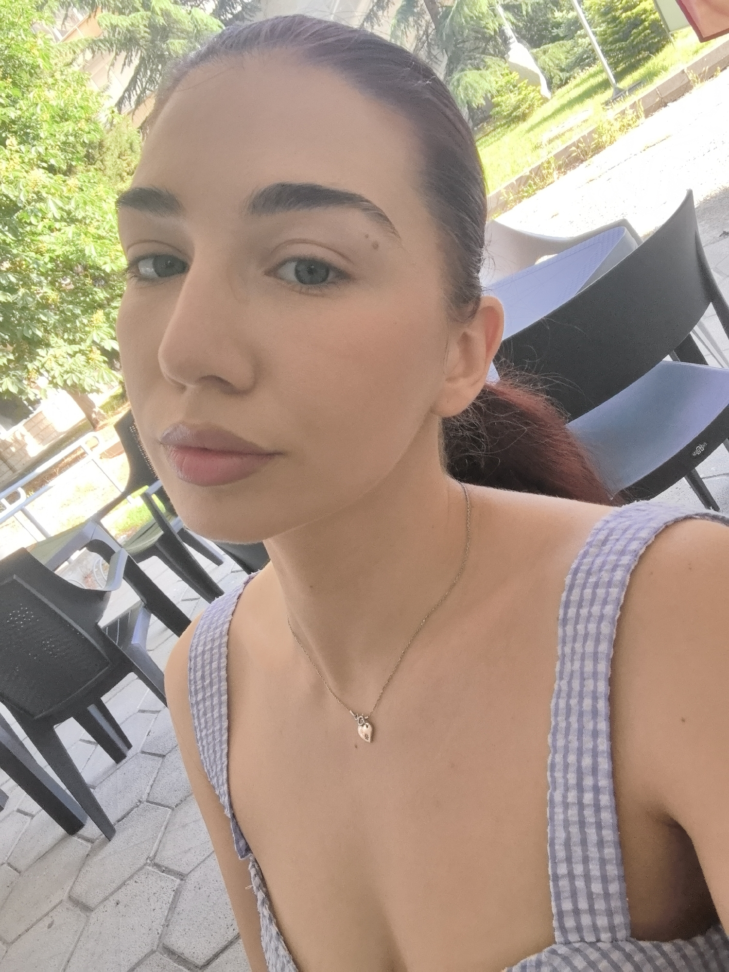 A young woman is posing for a selfie. She has fair skin, dark hair pulled back, and light-colored eyes. She is wearing a light-colored top with a gingham pattern, and a delicate necklace with a small heart pendant.
The background shows a patio area with dark gray chairs and tables. Trees and greenery are visible, suggesting an outdoor cafe or restaurant setting. The paved ground is patterned with gray stones. The photo appears to have been taken during the day. The location is Bogomilovo, Bulgaria.