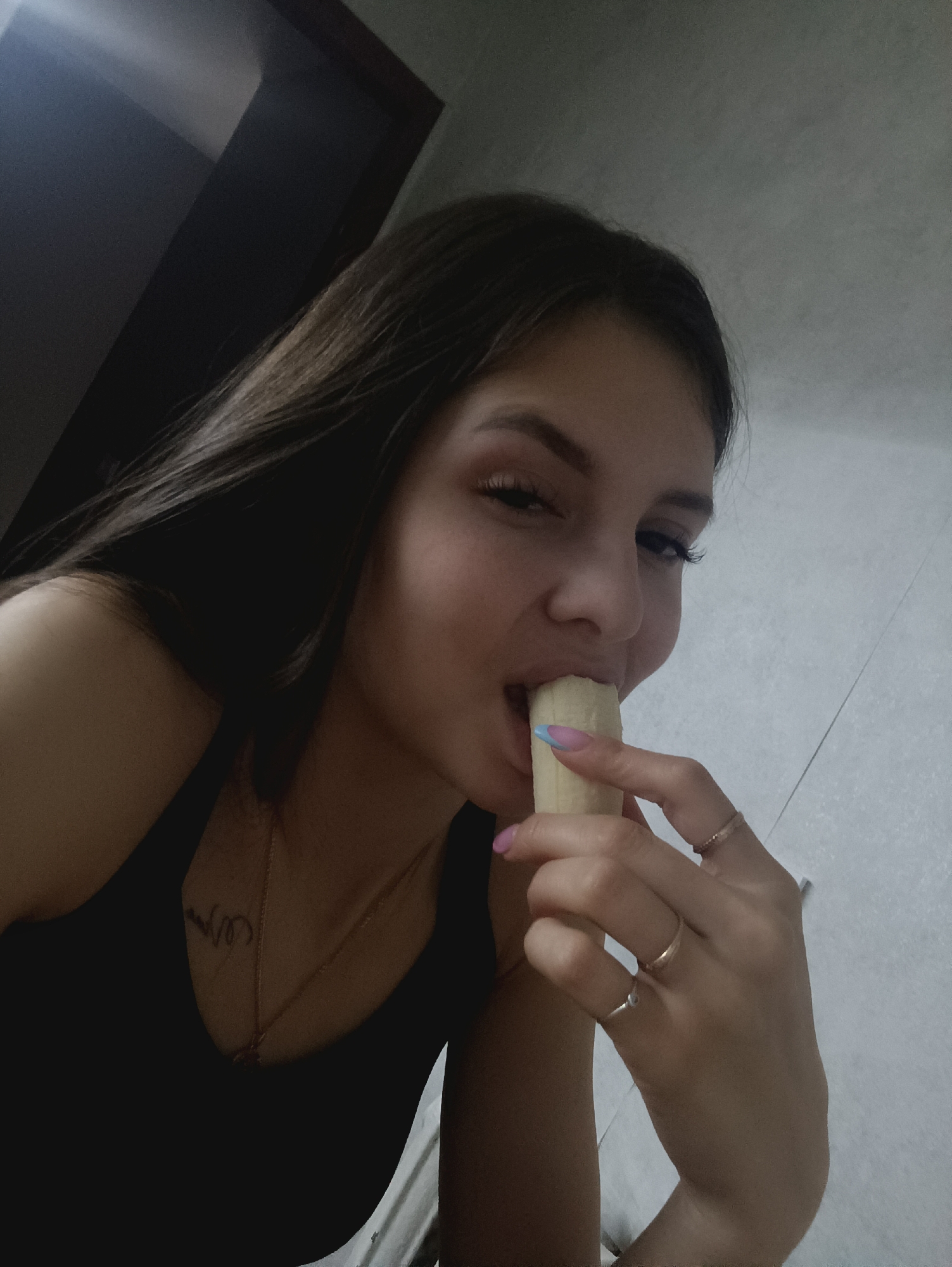 A young woman is holding and eating a peeled banana. She has long, dark hair and is wearing a black tank top and a necklace. She has a tattoo on her chest. She is wearing rings on her fingers and has pink and blue nail polish. The background is a plain white wall with a dark door frame in the upper left corner. The location is unknown.