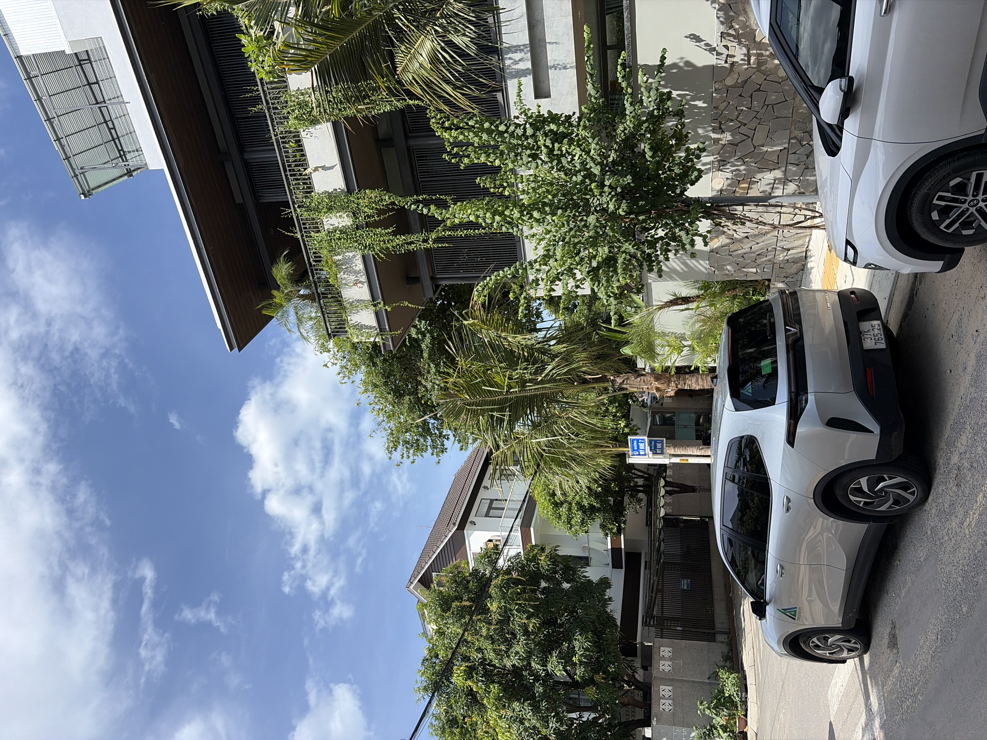 In a street scene in the My Khe area of Đà Nẵng, Vietnam, two silver SUVs are parked. The one on the left, a VINFAST VF e34, is positioned on the street, facing left. It has a green sticker on its rear window and license plate "37K 765.25". Partially visible on its side is the logo "THÀNH AN" with a green triangular design. The second SUV, a white car, is on the right, also parked on the street, facing left, with its front partially visible.
The backdrop features a multi-story building with modern architecture, characterized by large balconies adorned with hanging plants. Adjacent to it is a white and brown house with a pitched roof. Lush green trees, including palm trees, and foliage surround the buildings and line the street, creating a tropical ambiance. The sky above is a clear blue with scattered white clouds. A blue street sign with white text, reading "ĐƯỜNG MY KHÊ 2", is visible on a pole.