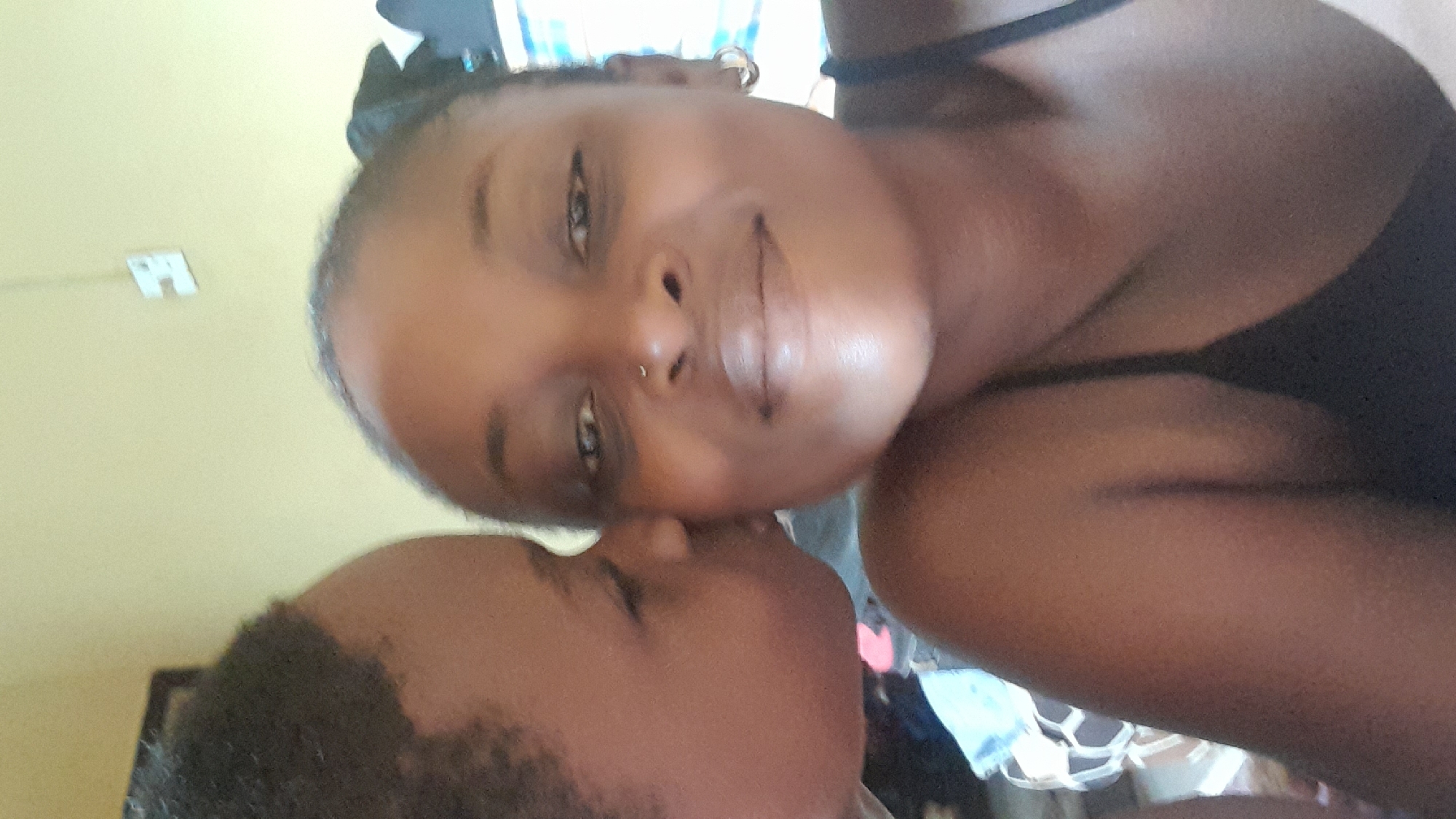 Two dark-skinned individuals, one appearing to be an adult female and the other a child with short hair, are close together. The child is positioned at the bottom of the frame, with the adult female above. It appears as though the child is kissing the adult female. The adult female is wearing a black top. The background is out of focus but appears to be a room, possibly in a residential setting in Ado, Nigeria. A light switch is visible on a yellowish wall. The angle of the photograph suggests it may have been taken as a selfie.