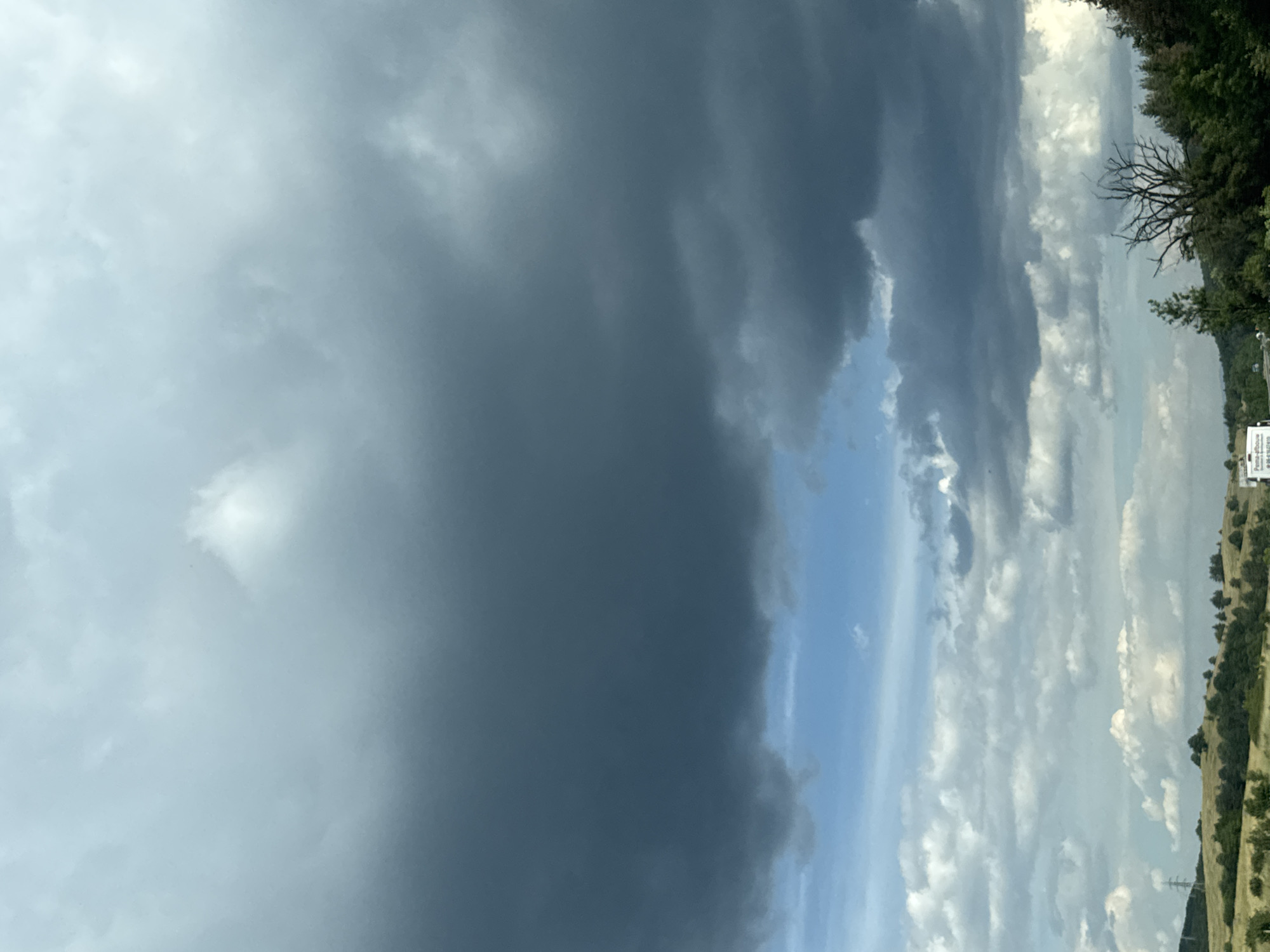 The image depicts a cloudy sky over a rural landscape, possibly in or near Rajana, Bulgaria. The upper portion of the frame is dominated by dark, ominous storm clouds, while the lower section shows a brighter sky with scattered cumulus clouds. The terrain below is hilly and covered with greenery, including trees and some dry grass. A power line tower is visible on the left side of the image, and a white truck with some text on it is partially visible at the bottom. The scene appears to be captured from a moving vehicle, suggested by the slight blur and the perspective.