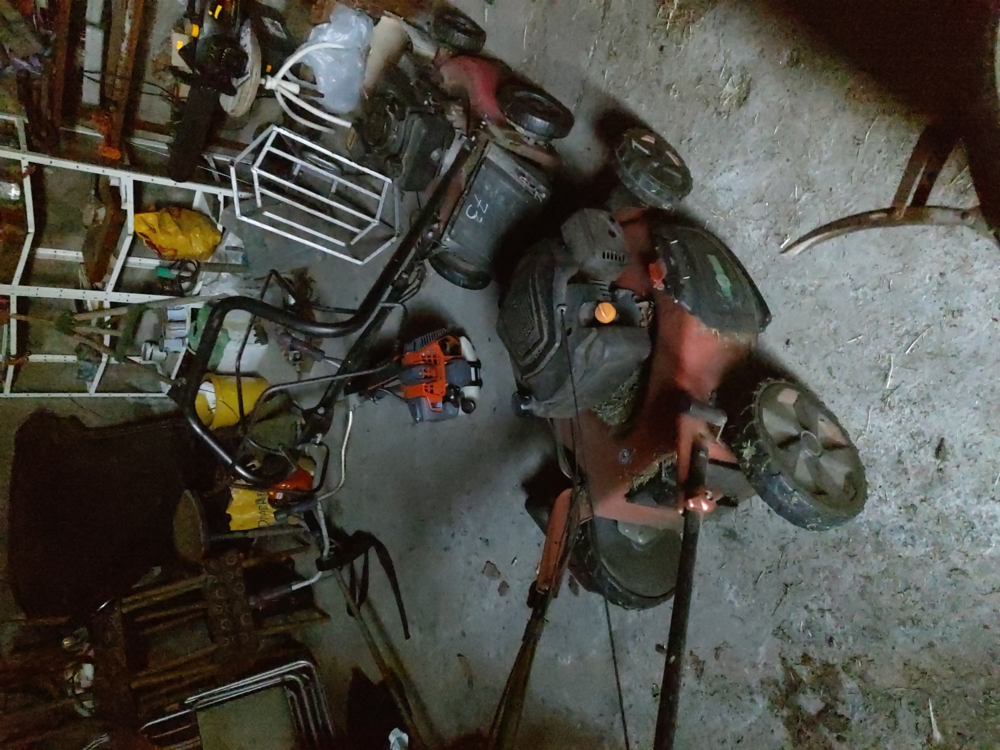 The image shows a collection of outdoor equipment, likely stored in a garage or shed. The primary subjects are two lawnmowers and a tiller. One lawnmower is predominantly red and black, with its wheels clearly visible. It appears to be a walk-behind model, tipped on its side. The other lawnmower is mostly obscured by shadows but seems to be a similar type. The tiller is orange and black, also on its side, with its tines visible.
Other objects in the scene include a yellow bucket, a white metal rack or cage, and various pieces of metal tubing and frames. There are also some plastic bags and what appears to be a yellow tarp or bag. The ground is a concrete or dirt floor, with some scattered debris and straw.
The lighting suggests an indoor setting, possibly dimly lit. There are no people visible in the image. The overall impression is one of storage or disarray, with the equipment not currently in use. The presence of lawnmowers and a tiller indicates a location where yard work is performed. The context provided, Kochmar, Bulgaria, suggests this could be a rural or suburban setting.
Visible text on the red lawnmower includes the number "75" on the engine housing. There is also some unreadable text on the yellow bag. The time of day and weather are not discernible from the image.