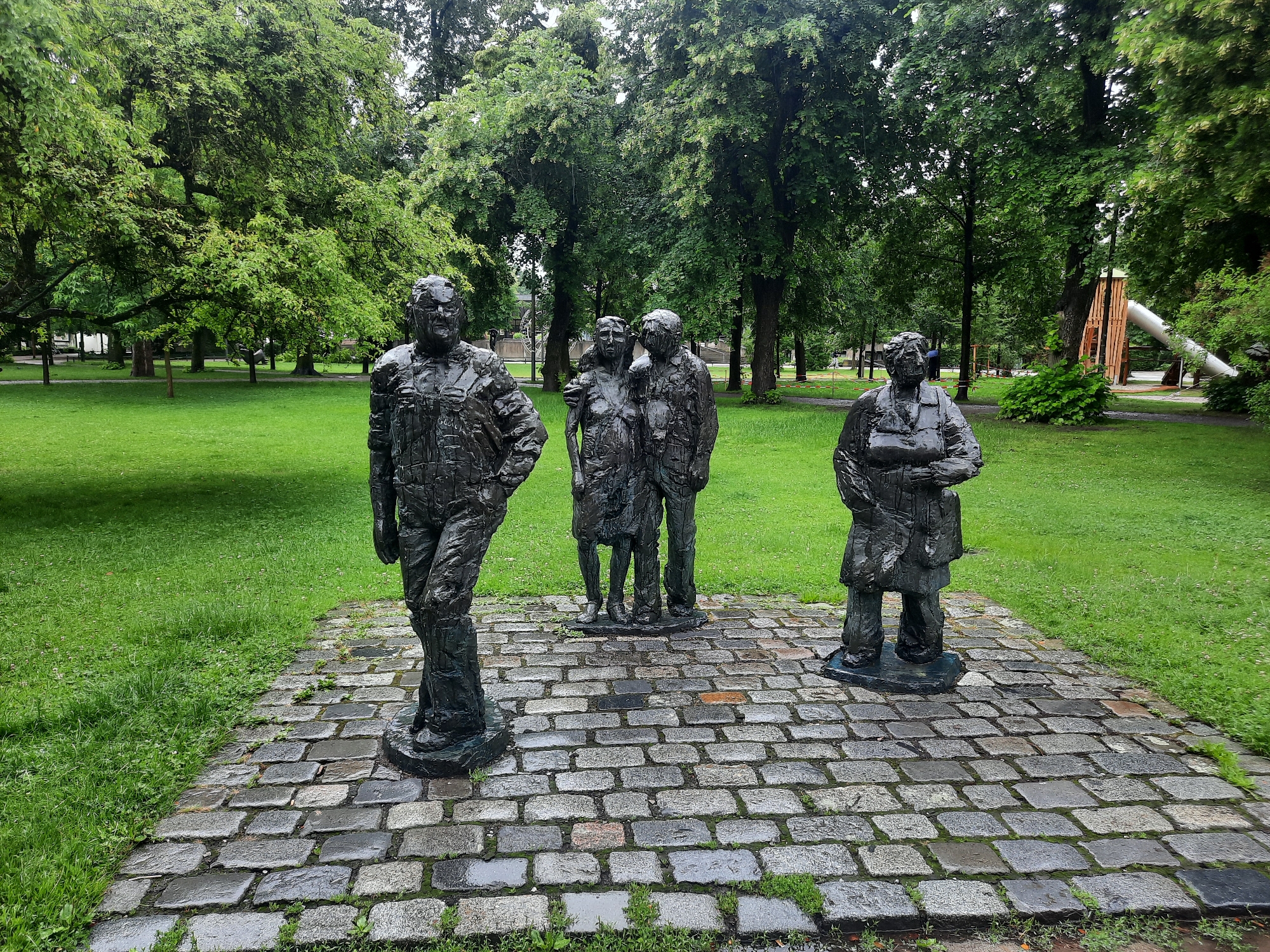 This image depicts a public park in Rosenheim, Germany, featuring a bronze sculpture of five figures standing on a cobblestone platform. The sculpture is located in a grassy area with mature trees in the background. To the right of the sculpture, a modern playground structure with a slide is visible. The sky appears overcast.