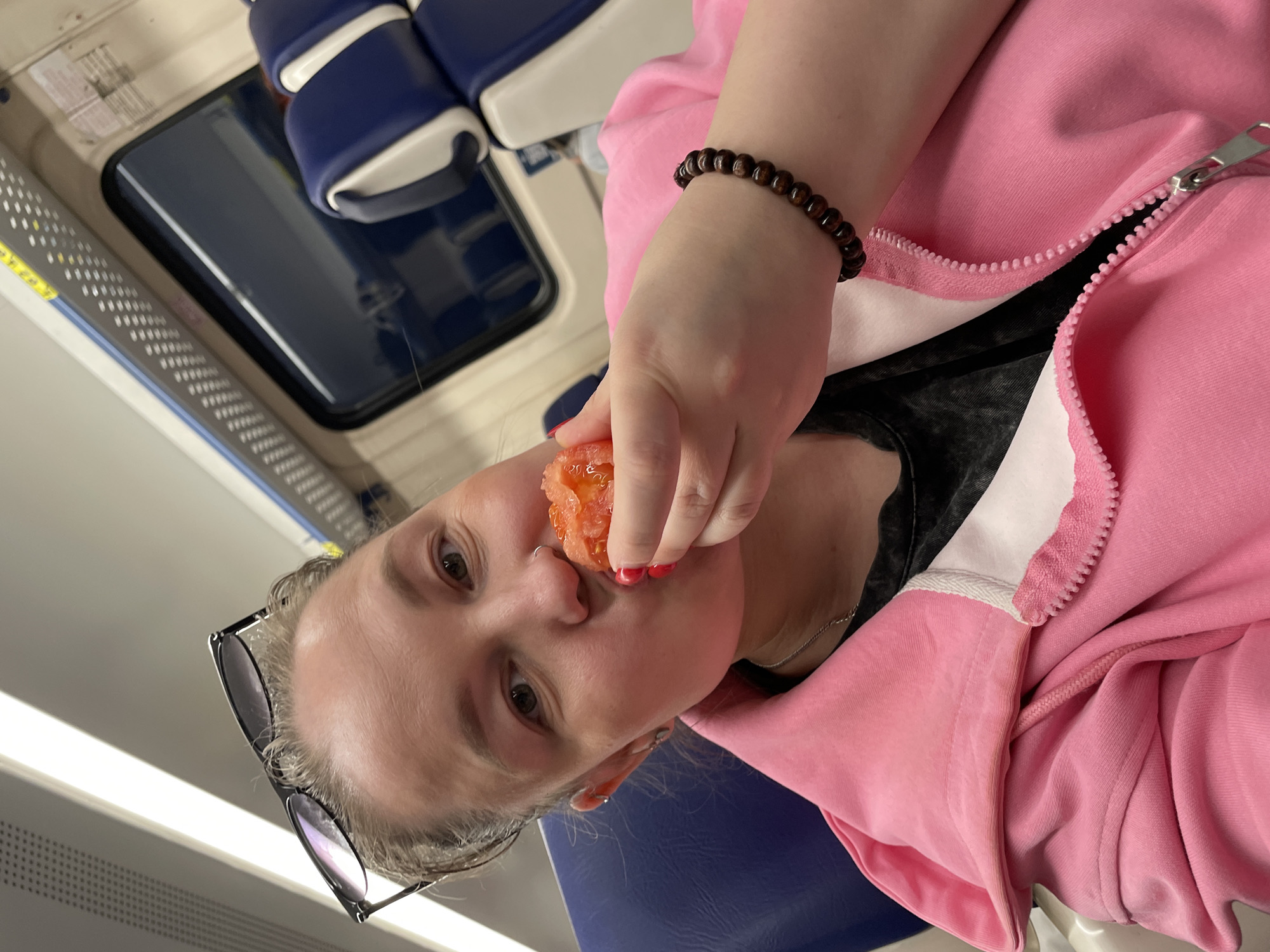 A person is holding a piece of food, likely a tomato, up to their mouth. They are wearing a pink zip-up jacket, a dark bracelet on their wrist, and black-framed glasses are positioned on their head. The background appears to be the interior of a train or bus with blue and white seats. The location is not explicitly apparent in the image; there is no clear indication of the city of Agryz, Russia.