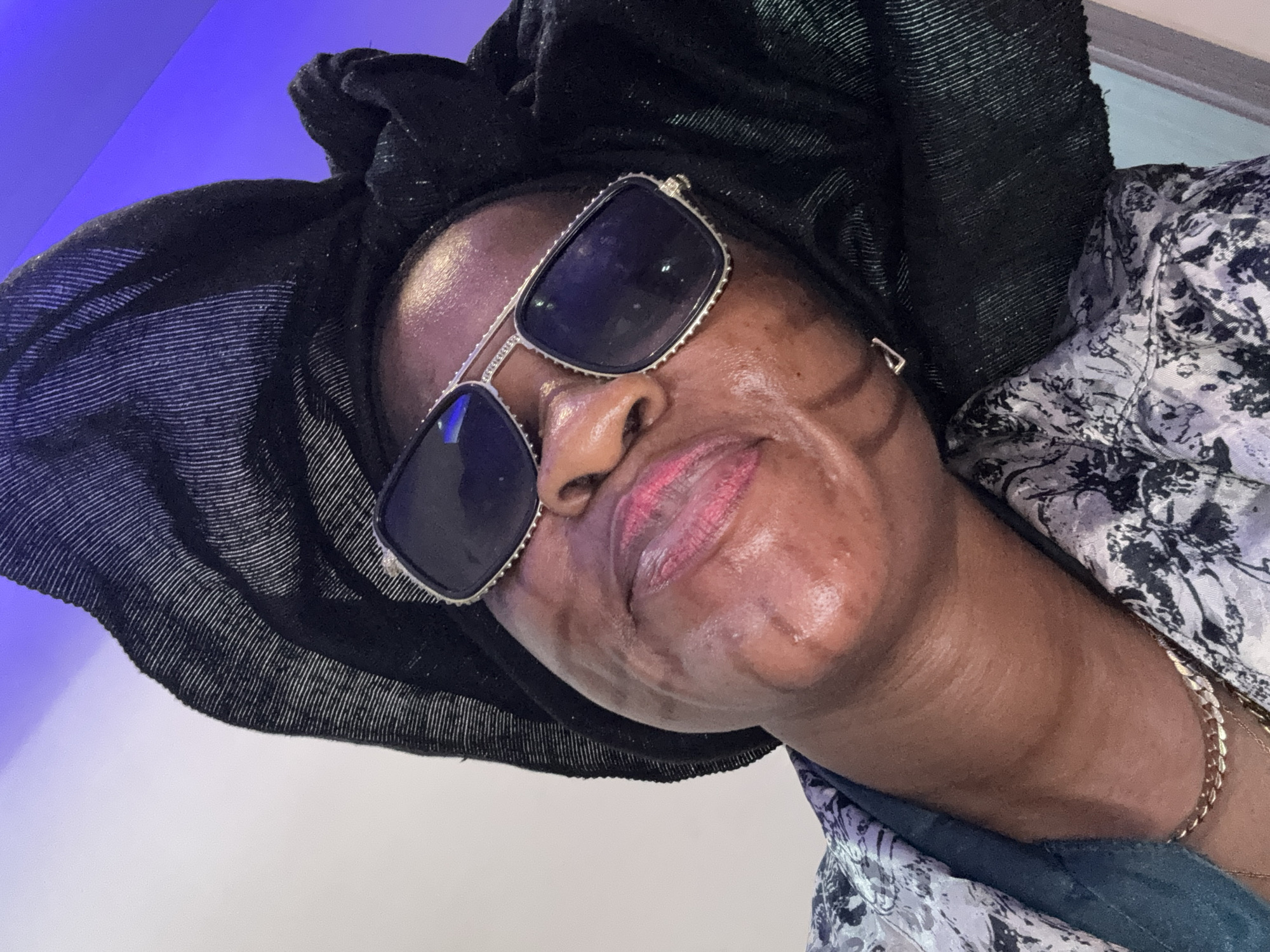 A medium-shot, low-angle, indoor photograph captures a woman wearing sunglasses and a black headwrap. The woman has her head tilted back slightly, and her face is illuminated by ambient light, creating highlights on her forehead and cheeks. She is wearing a patterned shirt with floral motifs in shades of gray and white over a dark green inner garment. A gold chain necklace is visible around her neck. The background features a white wall on the left, transitioning to a blue-lit area on the right. The photograph appears to be taken in a residential setting in Ajara, Nigeria.