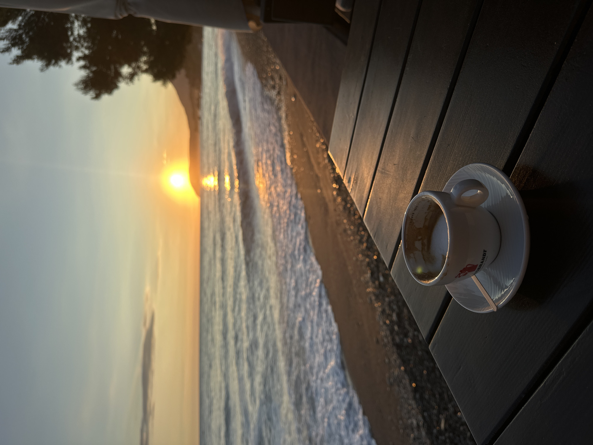 The image depicts a seascape at sunset in Šušanj, Montenegro, with a cup of coffee on a dark wooden table in the foreground. The sun is setting over the Adriatic Sea, its light reflecting off the water and the beach. Waves are visible along the shoreline. The cup is white, partially empty, and has a matching saucer. The table appears to be part of a seaside cafe or restaurant. No people are visible.