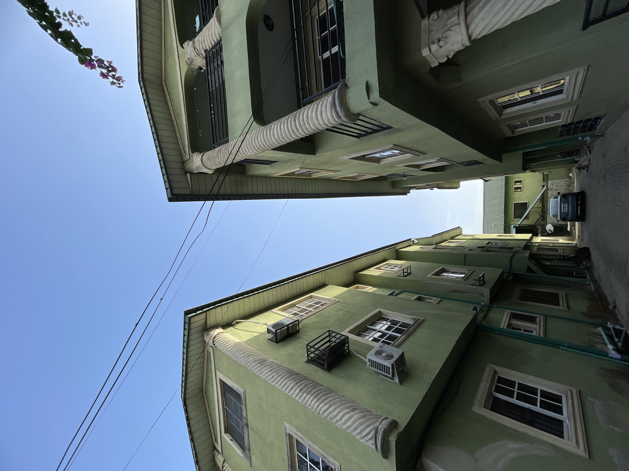 A ground-level, upward-angled perspective reveals a narrow passage between two multi-story, green-painted buildings in Ado, Nigeria. The clear blue sky is visible above the buildings. The buildings feature symmetrical window placement, decorative facade detailing that includes fluted column-like structures, and air conditioning units mounted on the exterior walls. A utility line stretches across the gap between the buildings, and a partial view of a flowering vine is visible at the upper left corner. Further down the passage, additional buildings are seen, including a light green corrugated structure and a parked van.
