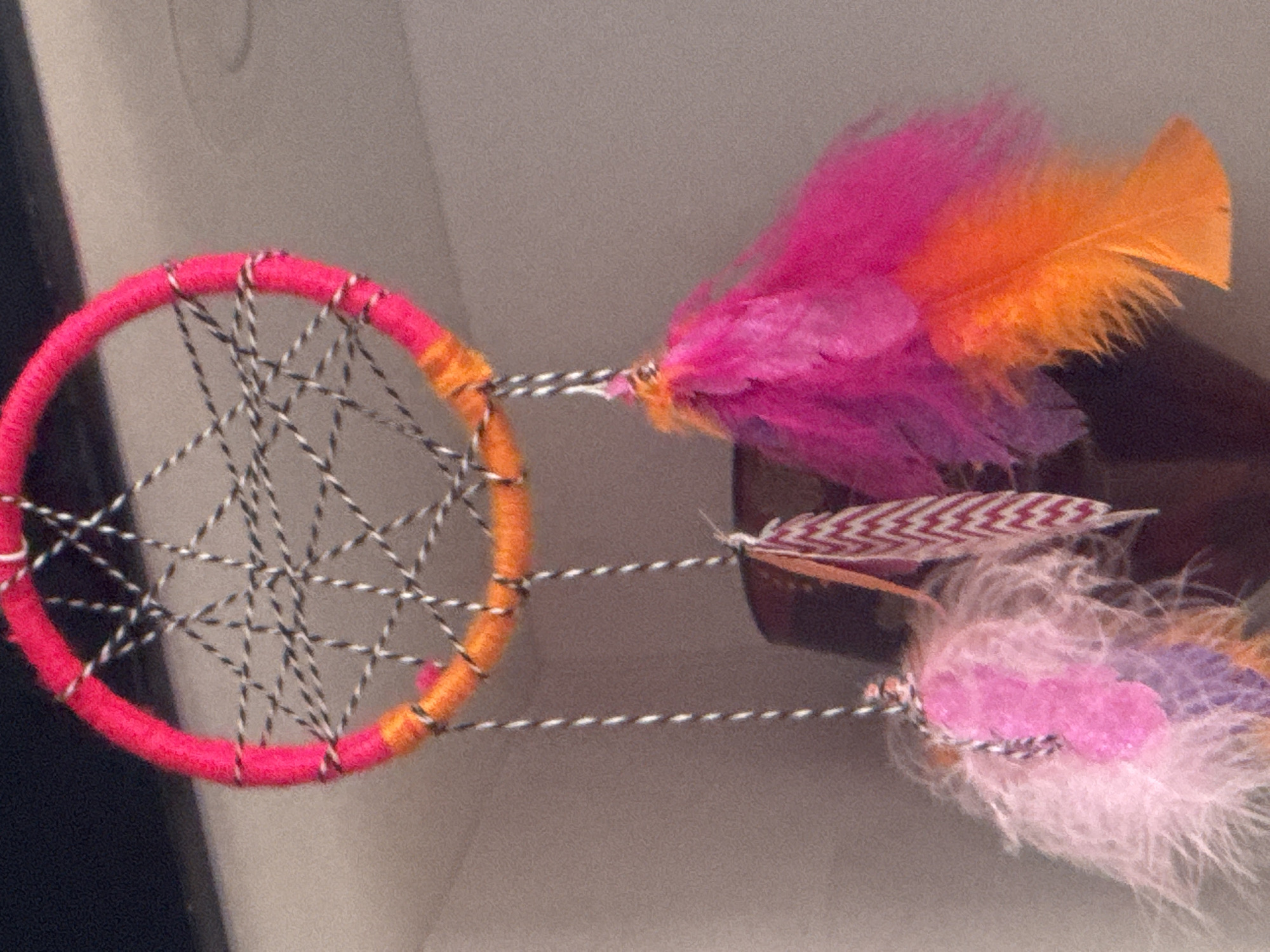 The image presents a colorful, handmade dreamcatcher-like ornament. The primary circular frame is wrapped in yarn, transitioning from a vibrant fuchsia pink on the left to a warm orange on the right. An intricate web is woven within the circle using black and white twisted string, forming a detailed pattern.
From the bottom of the ring, three separate strands of twisted black and white string extend downwards, each adorned with decorative elements. The uppermost strand features a cluster of fluffy feathers in bright fuchsia pink and vivid orange. The middle strand holds a single, flat feather or leaf-shaped piece, displaying a distinctive reddish-brown and white chevron or zigzag pattern. The lowest strand is decorated with multiple soft, fluffy feathers in shades of light pink, pale purple, and hints of light brown.
The background is a soft, indistinct light grey, suggesting an indoor setting, with a darker, blurred object partially visible on the right side. The overall aesthetic is cheerful and artisanal, highlighting the varying textures of the yarn, string, and feathers. There are no people or discernible activities taking place. No text is visible in the image. The lighting appears to be indoor, and the time of day is not evident. No specific location cues related to Tbilisi, Georgia, are present within the visual content.