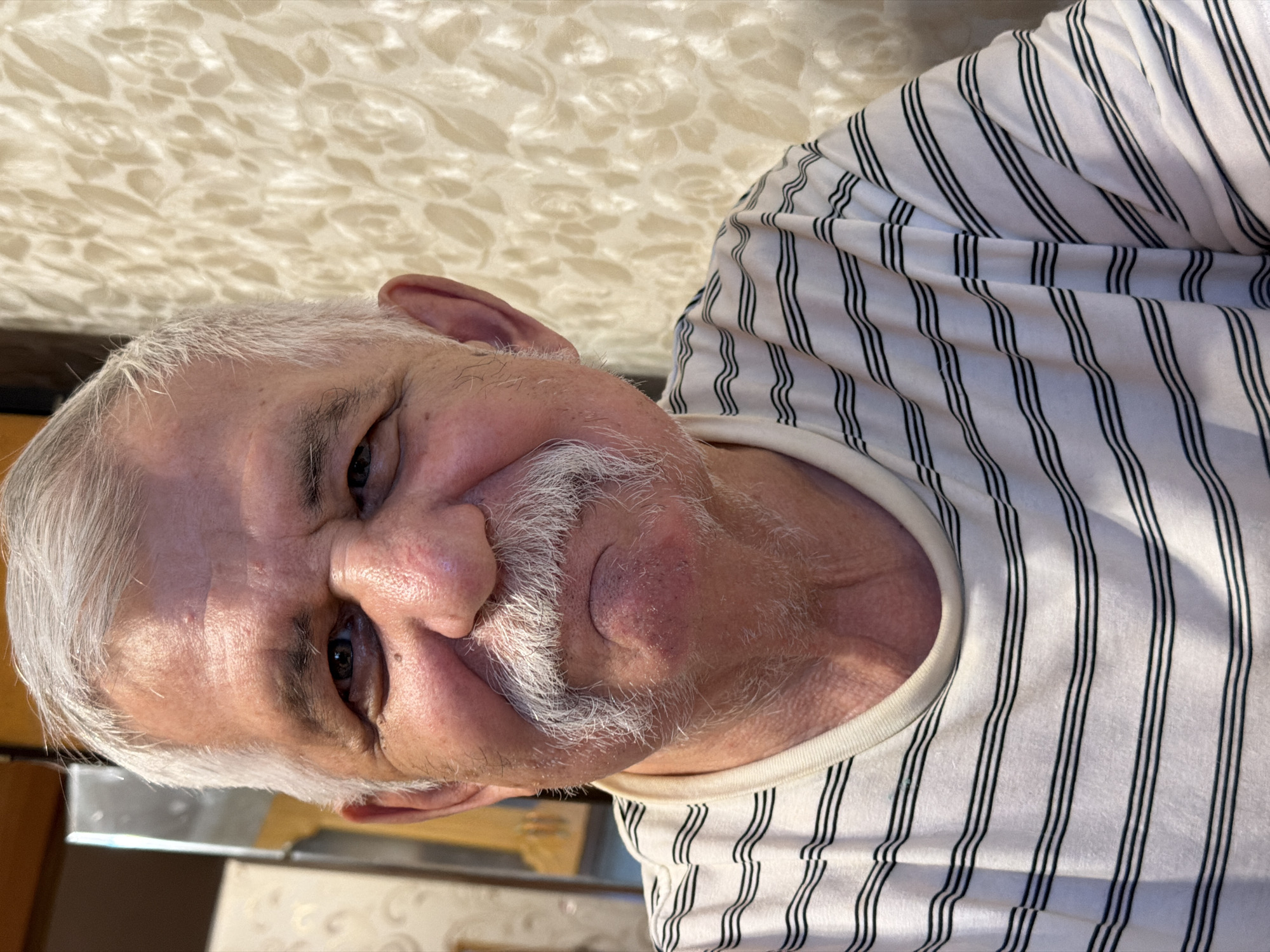 An older man with white hair and a white mustache is wearing a white and black striped shirt. The background is a beige and white textured surface. The photo is taken from a close-up, slightly angled perspective. The location in the city of Izhevsk, Russia, is not determinable from this image.