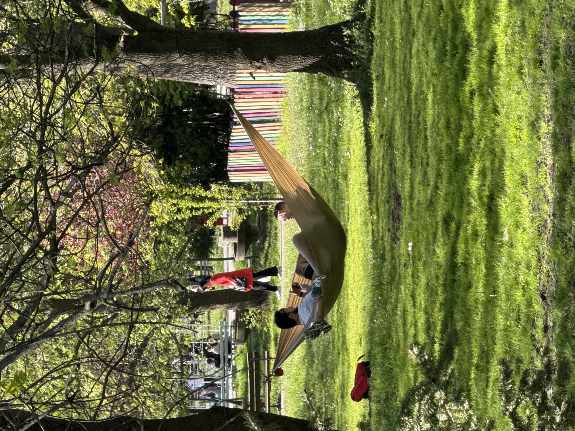 A sunny outdoor scene shows two people in a hammock slung between two trees on a grassy lawn. One person is sitting up, while the other is lying down. A red bag lies on the grass below the hammock. In the background, people are sitting at tables and walking, and a colorful structure resembling a xylophone is visible. The location is likely in Sofia, Bulgaria, based on architectural details and the presence of deciduous trees common to the region.