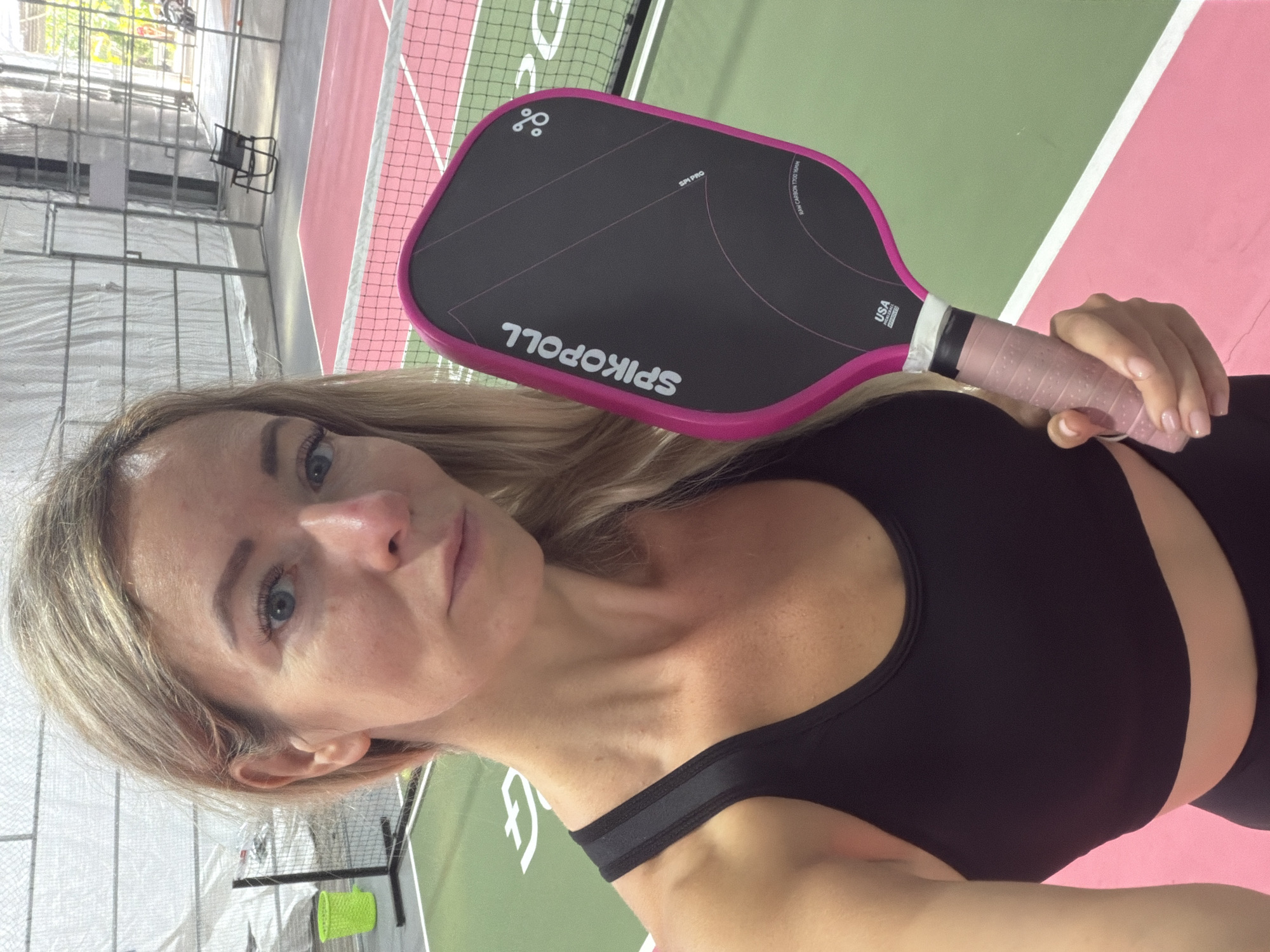 A blonde woman is holding a pink and black pickleball paddle. She is wearing a black sports bra and appears to be on a pickleball court. The court has green and pink surfaces, with a white line marking the boundary. In the background, there is a net, a green trash can, and a chair. The scene is indoors, with a corrugated metal roof and walls. The environment appears to be a recreational facility.