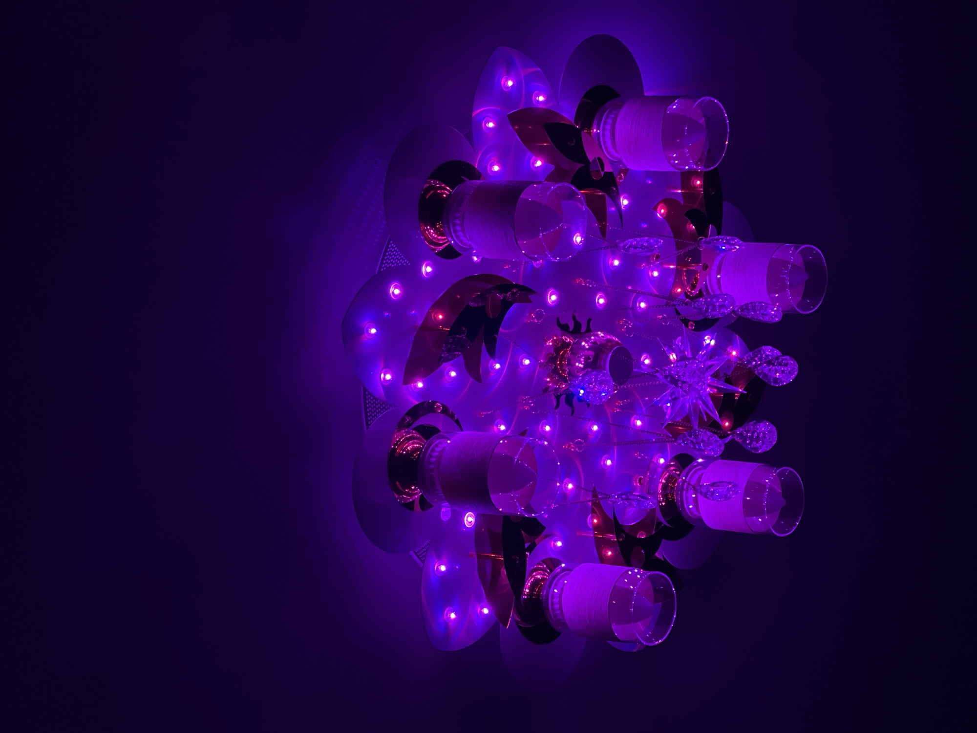 A complex light fixture is affixed to a dark surface, emitting a strong purple glow. The fixture consists of circular metal plates, glass tubes wrapped in white material, and ornamental, possibly crystalline, shapes. Small purple lights are scattered throughout the structure. The location and city are unknown.