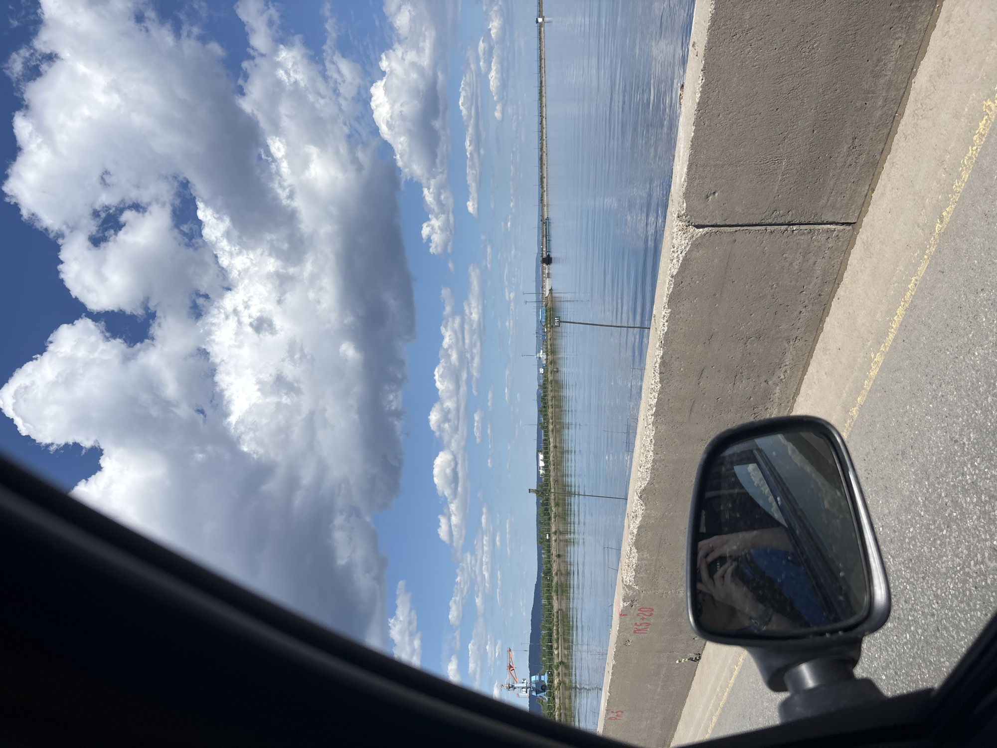 This image captures a panoramic view from the interior of a vehicle, looking out over a vast body of water, identified as the Votkinsk Reservoir in Chaikovsky, Russia. The shot is taken on a bright, sunny day with a clear blue sky dotted with numerous fluffy white and grey cumulus clouds.
In the immediate foreground, the dark grey frame of a car door is visible on the left, partially obscuring the view. Below this, a side mirror reflects a person, presumably the photographer, holding a phone to take the picture, along with parts of the car's interior and the road. The road surface, made of asphalt, is visible below the mirror, featuring a faint yellow line.
A prominent concrete barrier, likely a dam wall or retaining wall for the road, runs horizontally across the lower third of the image. Red markings, including "PK5" and "PK5+20", are stenciled onto this barrier. To the right of the concrete barrier, a black pole with a white rectangular sign reading "П11" extends vertically into the water.
The tranquil surface of the Votkinsk Reservoir occupies the midground, reflecting the blue sky and clouds. Numerous thin, dark poles or markers emerge from the water at various distances. Along the far bank, green vegetation, possibly trees and shrubs, is visible. On the left side of the far bank, a large blue and orange industrial crane stands out, flanked by other smaller structures and industrial elements.
Further into the background, a long, low-lying structure, potentially a causeway or part of the dam complex, stretches across the water. More industrial buildings and power lines are discernible along the distant horizon. The overall scene depicts a functional, industrial landscape alongside a large natural or man-made water body under a wide-open sky.