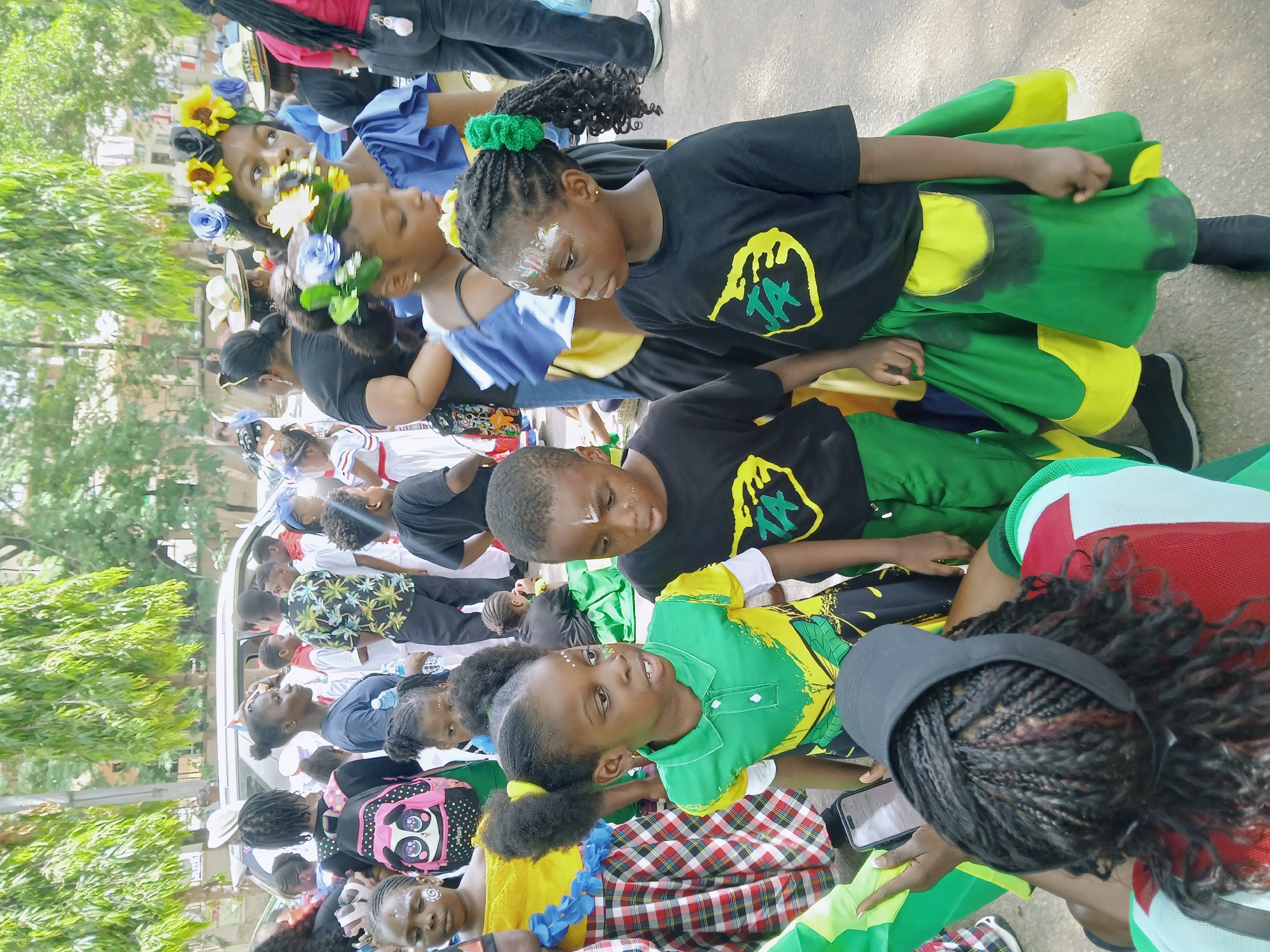 A medium-angle shot shows a group of people, mostly children, in an outdoor setting in what appears to be a street parade or festival. They are dressed in colorful outfits, including green, yellow, and red clothing, with some sporting face paint and flower headbands. Some of the children are wearing t-shirts with a distinctive yellow and green emblem. Trees are visible in the background, suggesting an urban environment. The location is inferred to be Abuja, Nigeria, based on the cultural attire and context of a street parade. People appear to be in motion, lining up or walking forward, and some are looking upwards. A person holding a smartphone is also visible in the lower right corner of the photo.