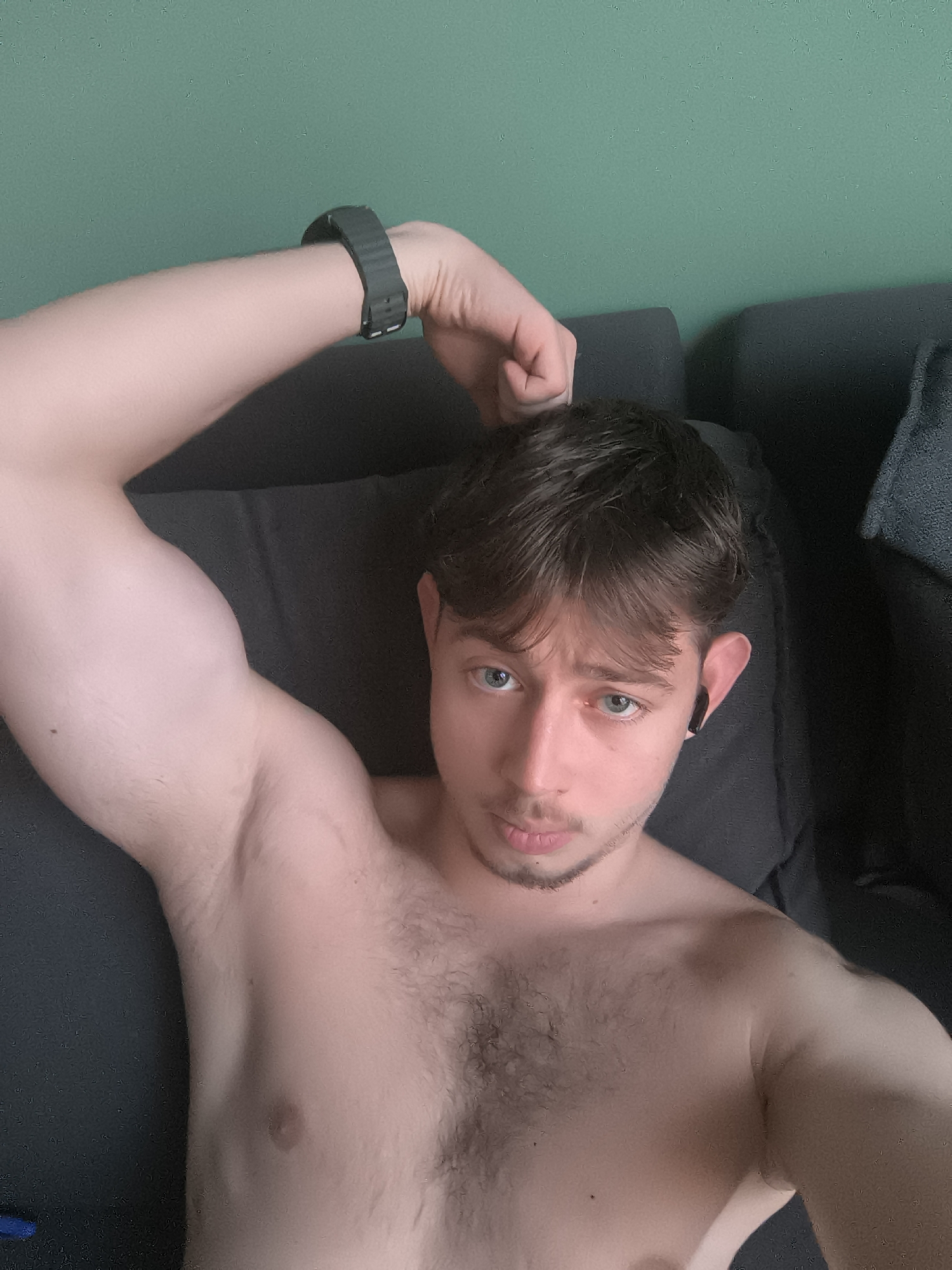 A shirtless young man with brown hair and blue eyes lies on a dark gray sofa. He is facing the camera with his left arm raised and bent, showing off his bicep. He is wearing a black fitness tracker on his left wrist and a black wireless earbud in his right ear. He has light facial hair. The background is a light green wall. The location cannot be determined as Sofia, Bulgaria from the image alone.