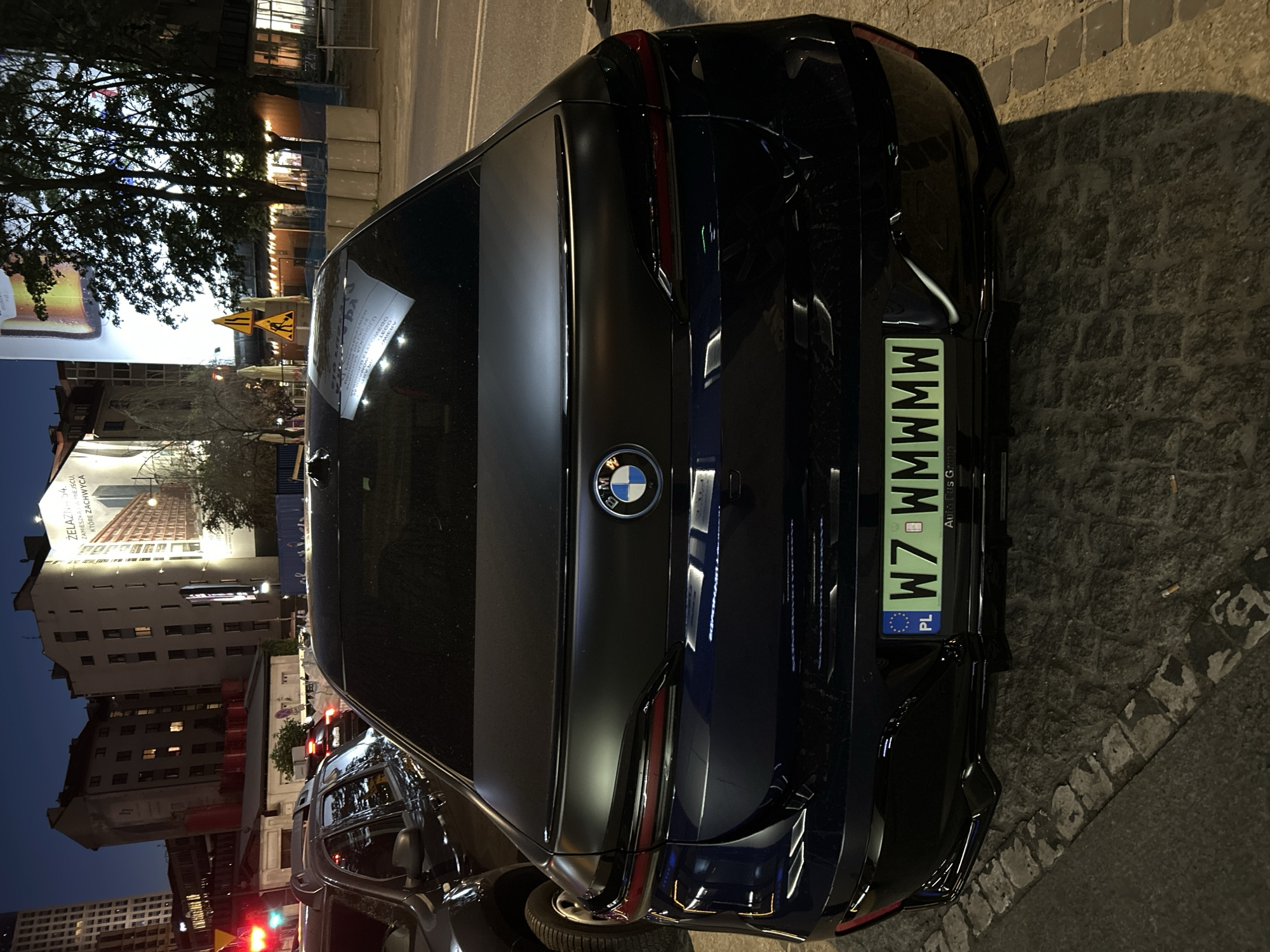 The image captures the rear view of a dark-colored BMW parked on a cobblestone street. The car is centered in the frame. The license plate reads "W7 WMWMW" and indicates that the car is registered in Poland ("PL" is visible on the plate).
In the background, city buildings and street lights illuminate the scene. A billboard is visible on one of the buildings, advertising real estate under the slogan: "Żelazna 54. Zamieszkaj w miejscu, które zachwyca" (Żelazna 54. Live in a place that delights). Street signs indicate the presence of road work or construction in the area. Other vehicles can be seen in the background, suggesting the car is parked on a busy street. Given the details, the location is plausibly Warsaw, Poland.