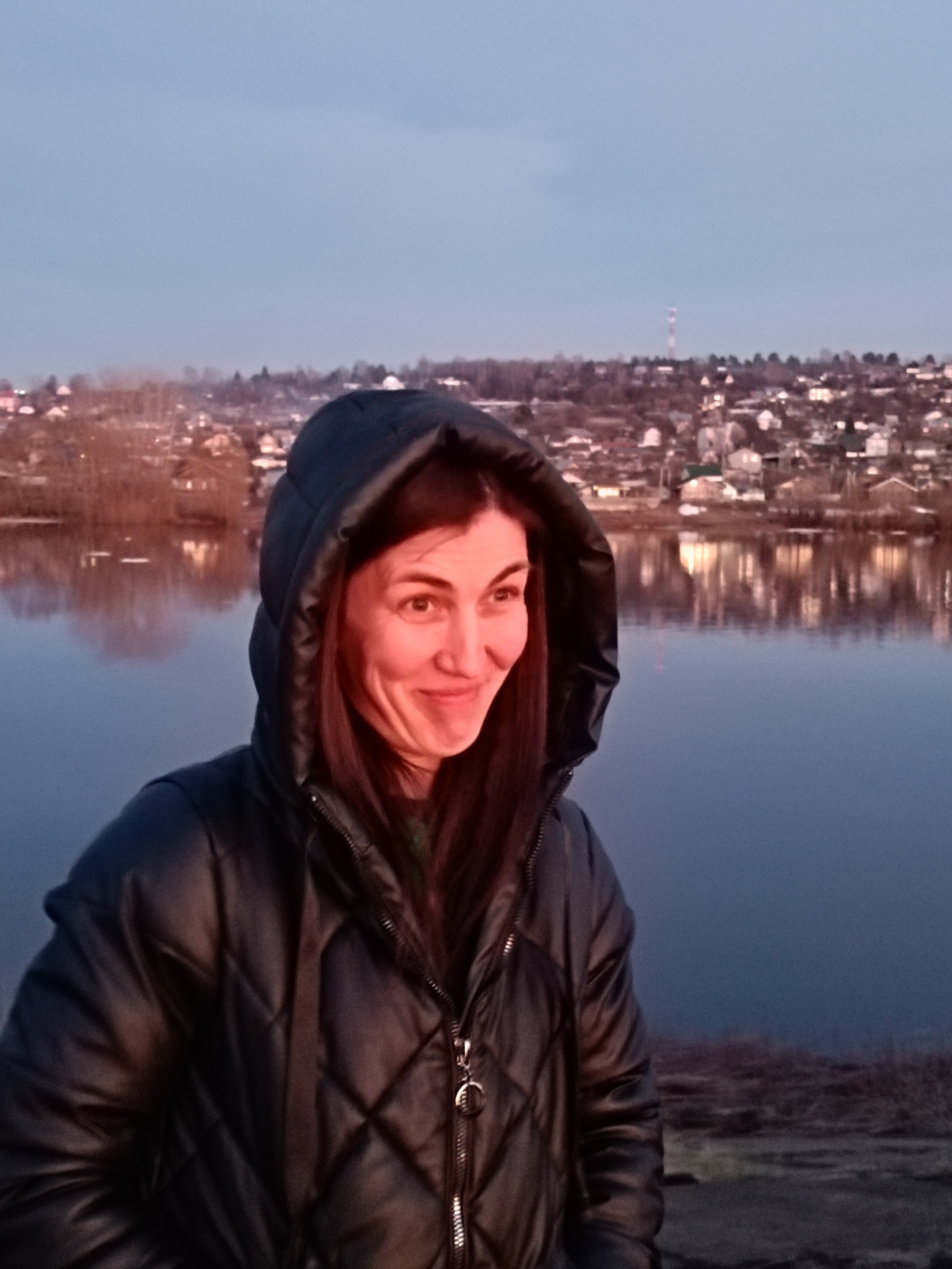 A close-up, chest-up shot features a woman outdoors during dusk or early evening in Chaikovsky, Russia. She has long, dark hair visible under a dark, possibly black, quilted puffer jacket with the hood up, framing her face. A warm, reddish light, likely from a low sun or artificial source, illuminates her face and parts of her jacket, contrasting with the cooler, dusky blue tones of the background. She displays a subtle, slightly mischievous or amused smile, looking off to the right of the frame. Behind her is a calm body of water, reflecting the scattered lights of a town nestled on the distant wooded bank. The town features numerous houses and buildings, some with illuminated windows, beneath a pale, hazy sky. Bare trees are visible along the distant shore. The scene evokes a quiet, somewhat chilly evening atmosphere. There is no visible text.