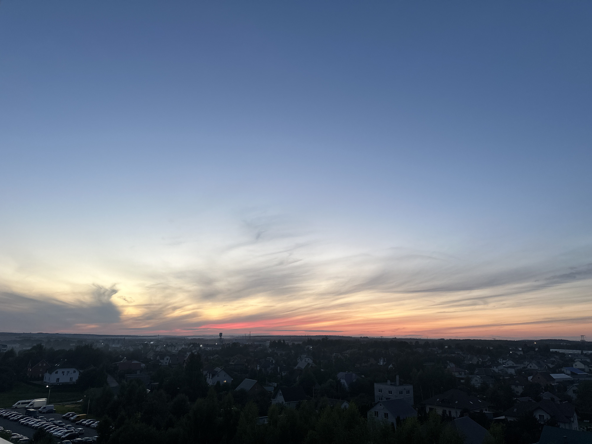The image is a wide-angle, high-angle view of a rural settlement at dusk, likely in the vicinity of Шчомысліцкі сельскі Савет, Belarus. The sky dominates the upper half of the frame, transitioning from a clear, deep blue at the zenith to softer hues of pale blue, pink, and orange near the horizon, indicative of sunset. Wispy clouds stretch horizontally across the sky, catching the last light of the day.
Below the sky lies a landscape dotted with numerous houses, mostly single-family dwellings with pitched roofs, nestled amongst dense green trees. The architectural style suggests a modern, suburban or rural development. In the foreground on the left, a large parking lot is filled with a multitude of cars, primarily silver and white, arranged in neat rows. A few vans are also visible. The lighting conditions are dim, with streetlights beginning to illuminate some of the houses and the parking lot. The overall impression is a quiet evening scene in a residential area.