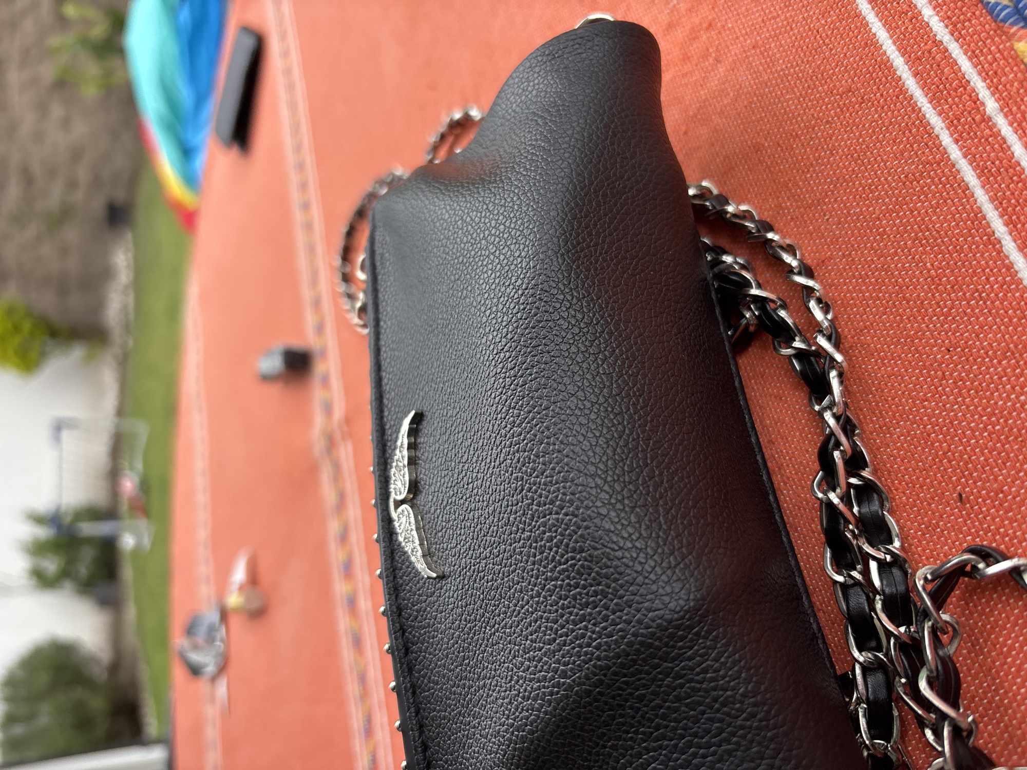 The image shows a close-up of a black leather handbag with a silver wing-shaped emblem. The bag has a silver chain strap intertwined with black leather. It rests on a table with an orange and white patterned tablecloth. The background is blurry, but reveals a green lawn, trees and a white wall. The location is most likely Puebla, Mexico due to the photo context.