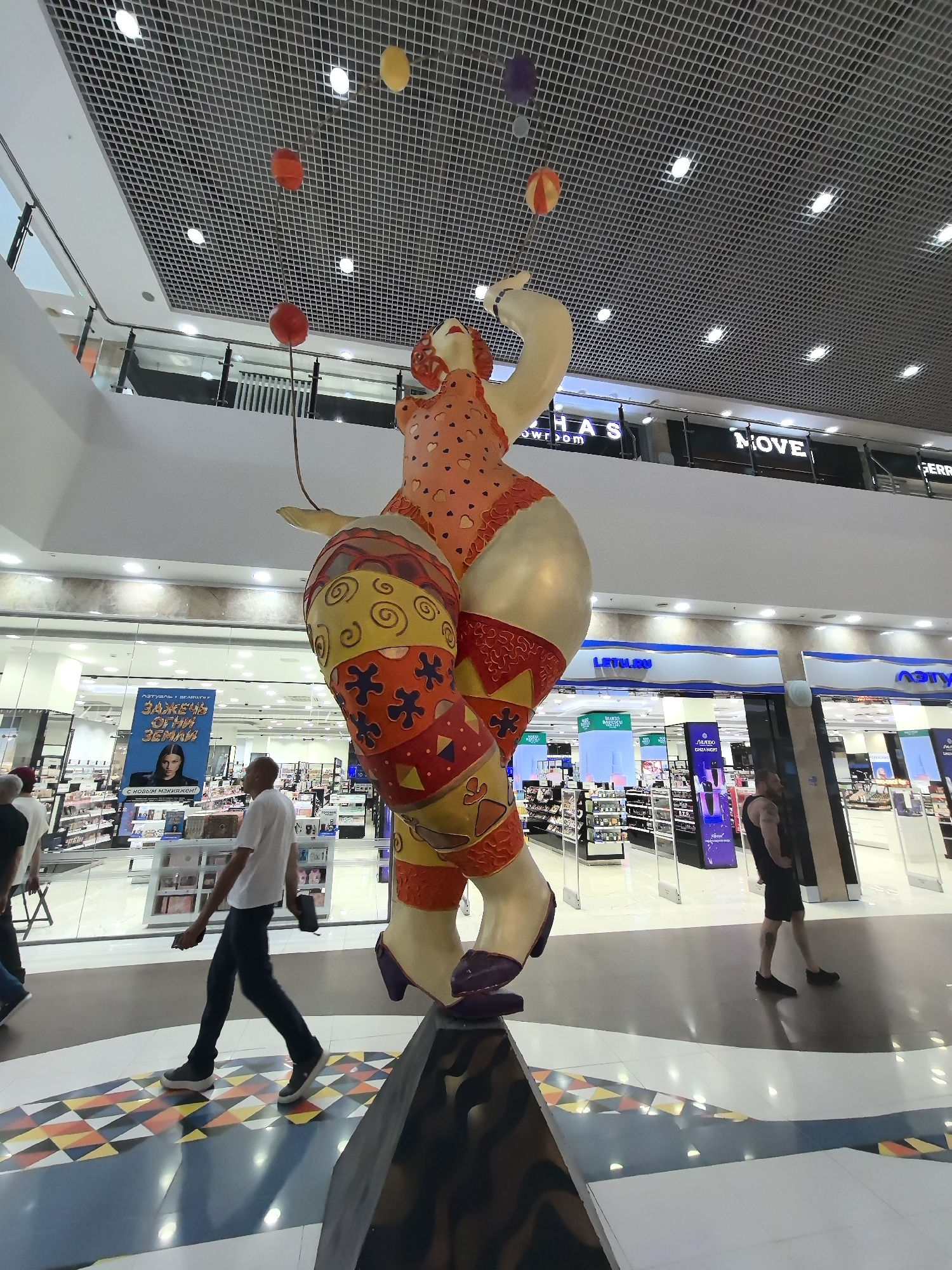 The image was taken inside a shopping mall. The main subject is a large, colorful sculpture of a woman juggling balls. The woman is depicted as stout with exaggerated features and is wearing a patterned outfit. She appears to be suspended in mid-air, with several red, yellow, and purple balls floating around her head, connected by thin strings. The background reveals the typical interior of a modern mall, with shops, displays, and shoppers walking by. Visible signs indicate commercial establishments, including "LETO.RU" and "Л'Этуаль" (L'Etoile), which are Russian beauty and cosmetics retailers. The floor has a geometric pattern, and the ceiling features a grid of lights. The overall scene depicts a public space with a prominent artistic installation. The mall is located in Nizhny Novgorod, Russia.