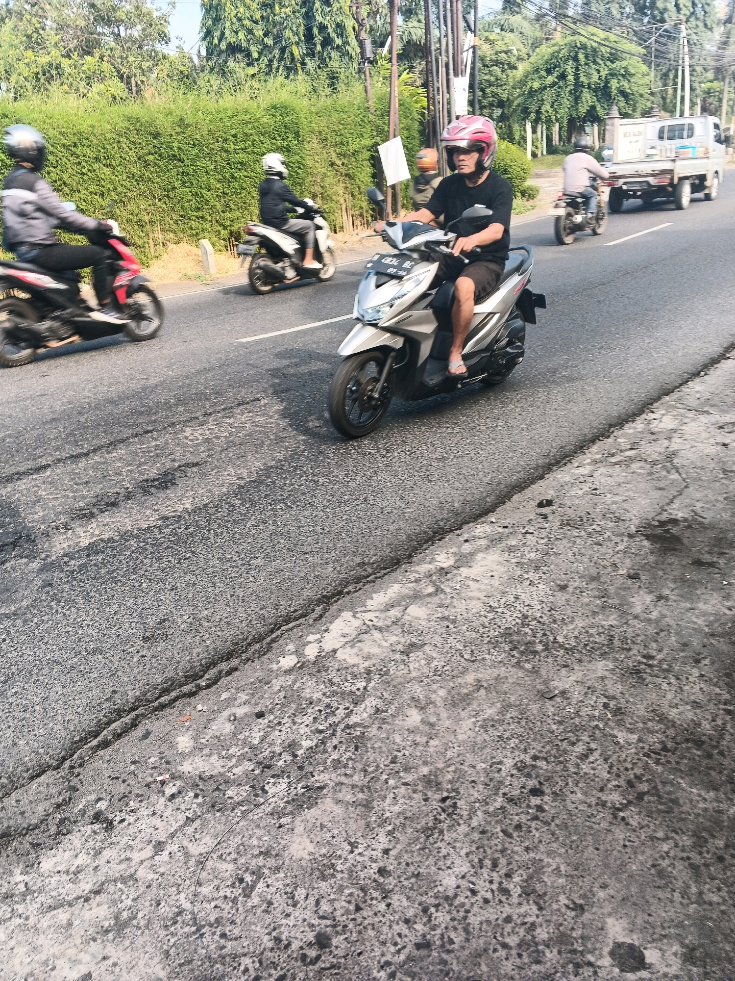 A man wearing a pink helmet and a black t-shirt rides a silver scooter on a road in Ungaran, Indonesia. He is wearing shorts and flip-flops. The scooter has a license plate that reads "1834 BC". In the background, another person rides a white scooter and a white truck is visible. The road is paved and has some cracks. There are trees and a hedge on the side of the road. The image appears to be taken during the daytime.