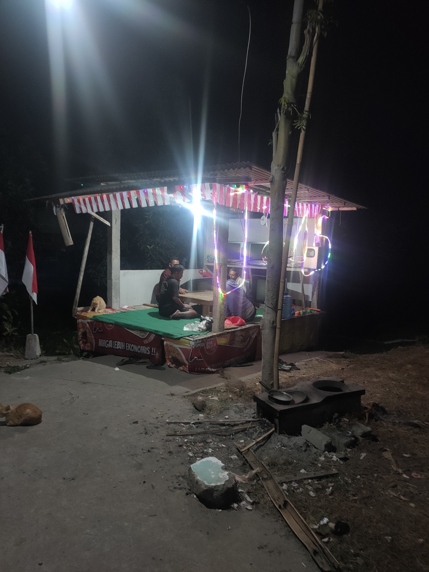 The image depicts an outdoor scene at night, likely in Kanigoro, Indonesia, with a small, makeshift food stall illuminated by string lights and a bright overhead light. The stall is decorated with Indonesian flags. Two individuals are seated inside the stall, appearing to be conversing or working. A cat is visible near the stall, and another cat is lying on the ground in the foreground. In the background, there is a traditional stove with a pan on it. The ground around the stall is uneven and appears to be a mixture of dirt and debris. The overall atmosphere is that of a humble, local establishment operating after dark.