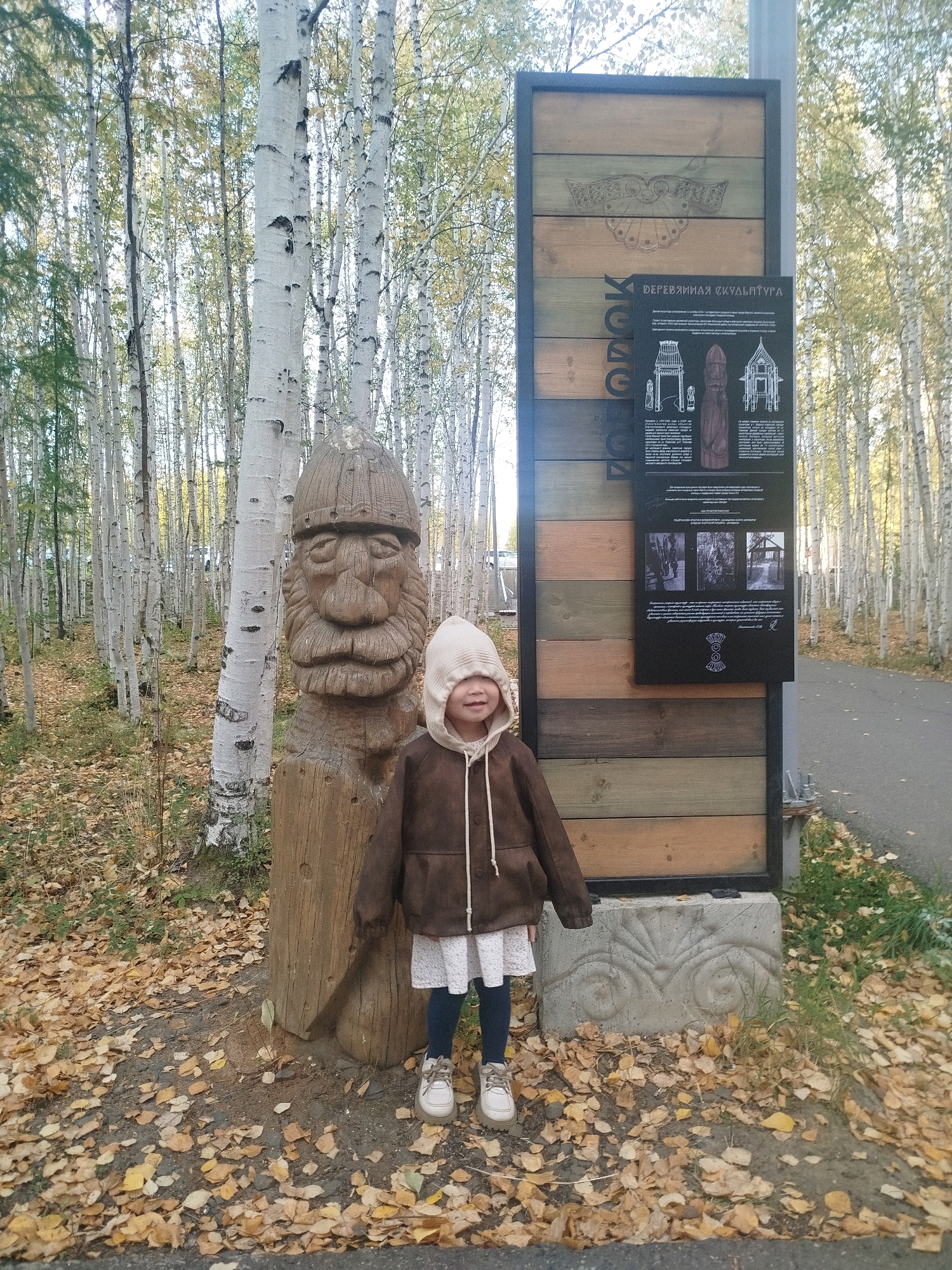 In Mirny, Russia, a young girl stands next to a wooden sculpture of a bearded man's head. The girl, wearing a hooded brown jacket, white dress, and light-colored shoes, is positioned slightly to the right of the sculpture. Behind them is a tall information board made of wooden planks, featuring text and images related to "Wooden Sculpture". The scene is set within a birch forest during autumn, as indicated by the fallen yellow leaves covering the ground and the trees with white bark and sparse foliage. A paved path runs alongside the forest on the right side of the image.