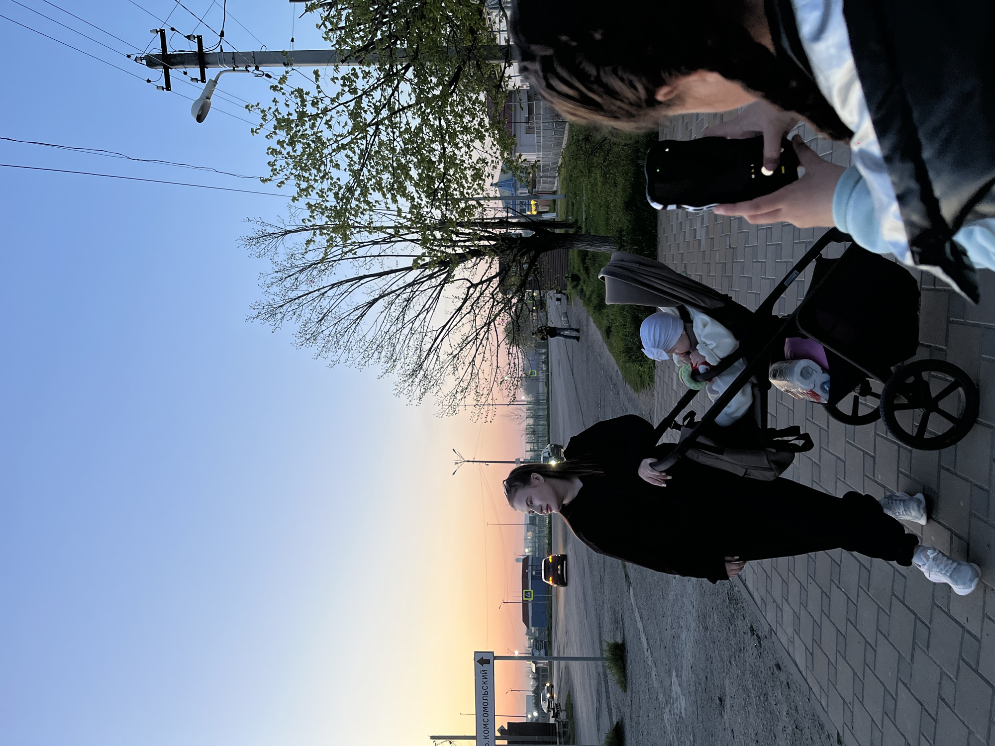 An outdoor scene captured at dusk or sunset in Novopokrovskaya, Russia, shows a group of people on a paved sidewalk next to a road. The image is taken from a high angle, looking down and slightly forward.
In the foreground, a person's head and hands holding a smartphone are visible at the top right, suggesting they are capturing the scene. Ahead of them, a woman in a black long-sleeved top and black pants, wearing white sneakers, pushes a dark-colored baby stroller. Her left hand is on the stroller handle, and her right hand is near her side. Inside the stroller, a baby wearing a white cap is visible, holding a red toy.
The sky transitions from a light blue at the top to a warm orange and yellow glow at the horizon, indicating late evening. On the left side of the frame, a tall utility pole with a street lamp and power lines extends into the sky. Several trees with sparse foliage and bare branches line the sidewalk, particularly prominent on the left.
The paved sidewalk gives way to a road on the right, where a car with illuminated headlights is visible in the distance, driving towards the sunset. Another vehicle is also partially seen further down the road. Buildings and structures line the horizon.
A vertical road sign in the lower left features an upward-pointing arrow and reads "пр. КОМСОМОЛЬСКИЙ" (Komsomolsky Prospekt/Ave.), confirming the urban setting and providing a specific street name within the given location context. The overall atmosphere is calm and serene, characteristic of the transition from day to night.