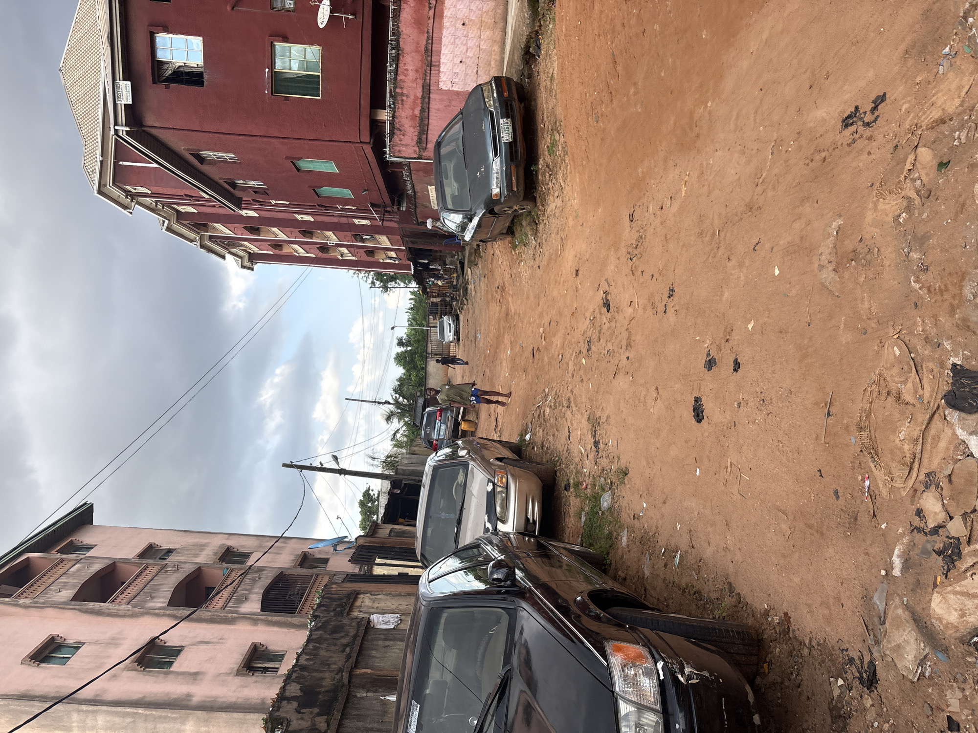 This is a ground-level, medium shot taken on a dusty, unpaved street in Onitsha, Nigeria. The street is lined with buildings, one on the left with a pale pink facade and balconies, and another on the right painted a deep red, with a white-tiled roof. Several cars are parked along the street, including a black sedan on the left, a light gold sedan in the middle, and a dark sedan with a license plate starting with "K10" parked on the right. Two young men are walking on the street; one, wearing a green t-shirt and shorts, is walking away from the camera towards the right, while the other, wearing a dark shirt and blue shorts, walks towards the left. The ground is a reddish-brown dirt, littered with debris. The sky is cloudy and overcast.