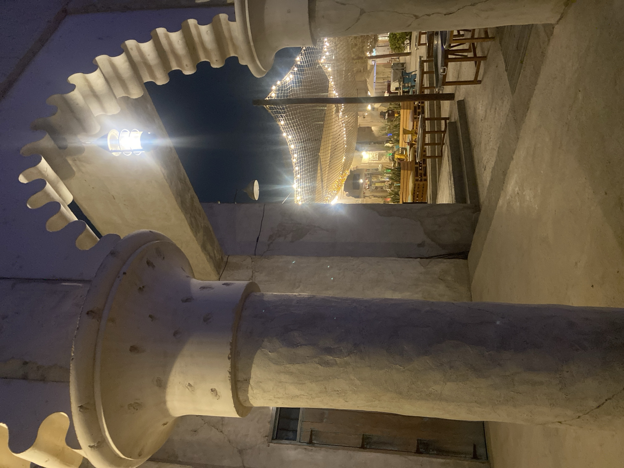 The image depicts an outdoor dining area at night in Deira, United Arab Emirates. The foreground features a large, rough-textured concrete pillar on the left and a similar architectural element on the right, framing an arched entryway. Beyond the archway, strings of fairy lights are suspended over an area with tables and chairs, suggesting a marketplace or restaurant setting. A rustic wooden door is visible on the far left of the frame. The overall atmosphere is illuminated by artificial lighting, casting a warm glow on the scene.