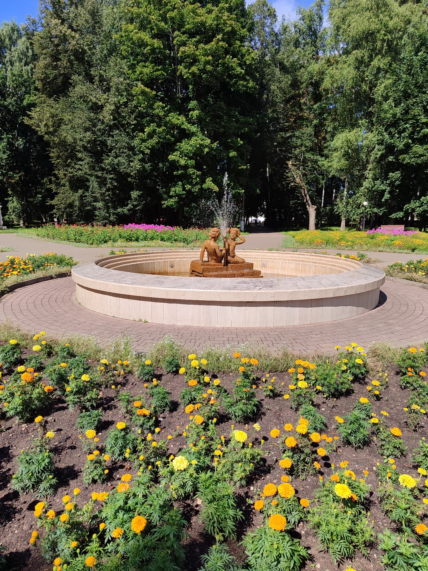 An eye-level medium shot captures a stone fountain with two cherubic statues at its center, located in a sunlit park. The fountain is surrounded by a circular brick pathway, which is itself encircled by a bed of vibrant yellow and orange marigolds. Beyond the flowerbed, a wider brick pathway curves through the park, leading towards a cluster of lush green trees. In the background, more trees and a few scattered pink flowers add depth to the scene. The sky above is a clear, bright blue.