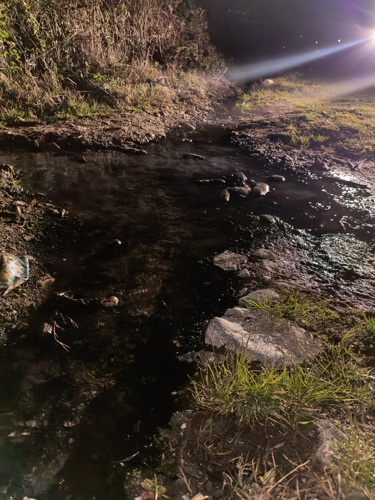 A dark, shallow stream flows through an area of dirt and vegetation under the light of a bright source, possibly a streetlight. The water appears murky and dark, and rocks are visible within the stream. The stream bank consists of dirt and patches of grass, with taller vegetation visible in the background. A piece of trash is visible on the left bank of the stream. The location is labeled as Unknown, Unknown. There are no people visible.