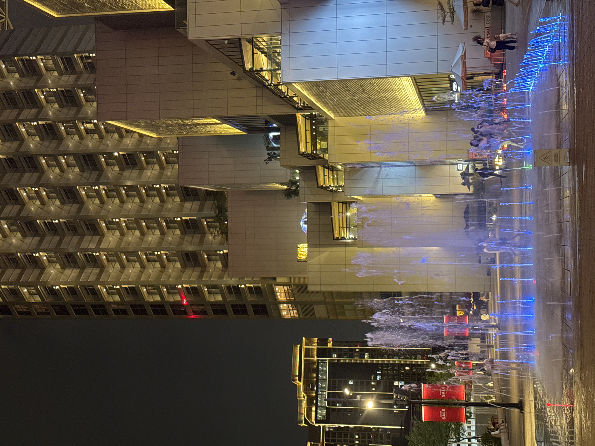 The image depicts a nighttime scene at Vincom Center Metropolis in Hanoi, Vietnam. A modern architectural complex with illuminated buildings and balconies is visible in the background. In the foreground, a water fountain with colorful lights creates a dynamic display. Several people are present, some interacting with the fountain, while others walk or sit in the vicinity. Red banners with the word "SALE" are hung on lampposts to the left of the scene. The overall atmosphere is lively and urban.