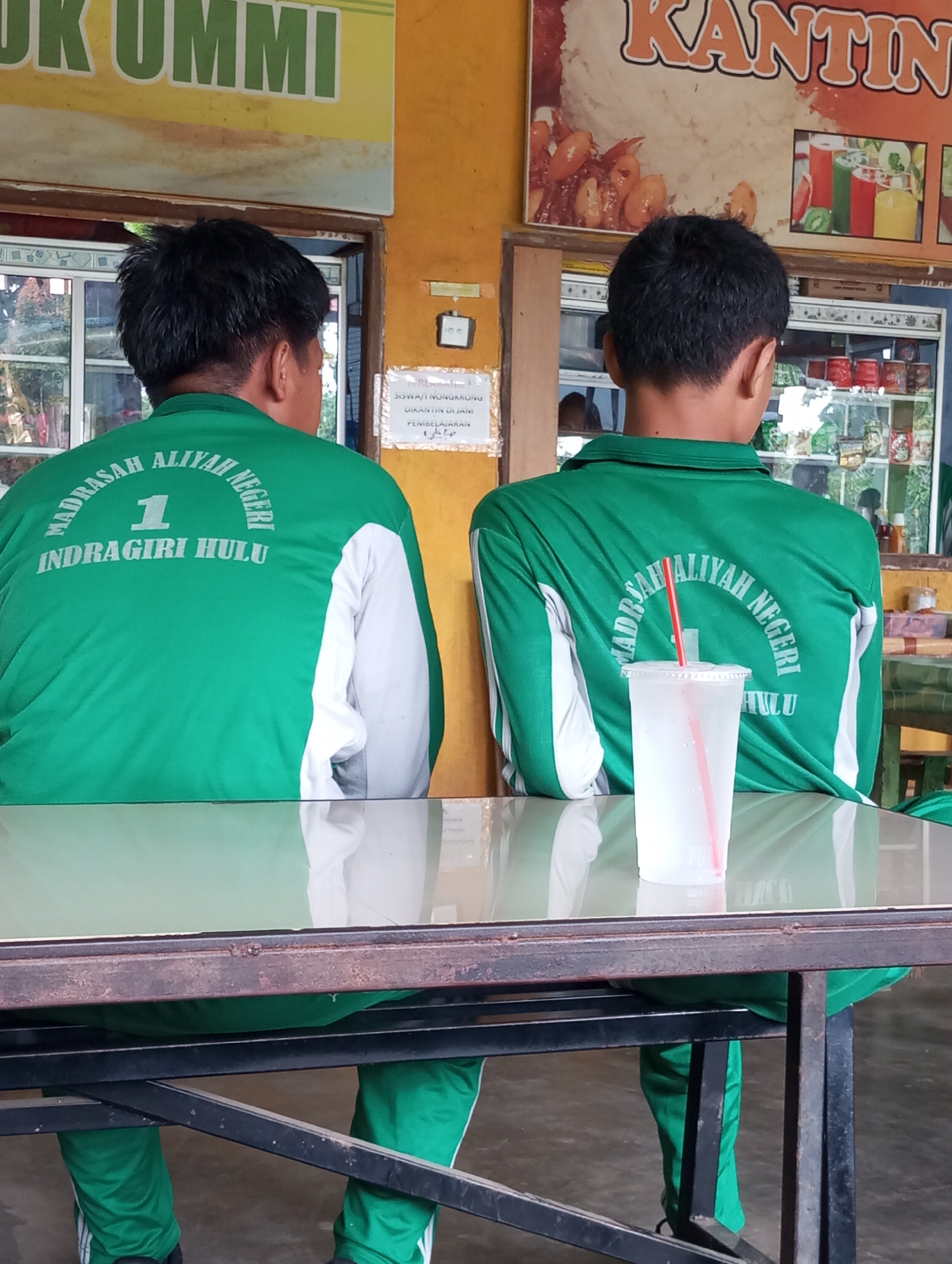 Two individuals, viewed from the back, are seated at a table in what appears to be a cafeteria or canteen in Pematang Reba, Indonesia. Both are wearing green tracksuits with white accents on the sleeves. The tracksuits have white lettering on the back. One reads "MADRASAH ALIYAH NEGERI 1 INDRAGIRI HULU" and the other shows "MADRASAH ALIYAH NEGERI". A clear plastic cup with a red straw, filled with a light-colored liquid, sits on the table in front of the individual on the right. The background reveals a yellow wall with signage, including "KANTIN" which translates to "Canteen", and another sign with green lettering that is partially visible. Shelves stocked with various items are also visible behind the individuals. The overall setting suggests a casual environment, possibly associated with a school or educational institution given the inscriptions on the tracksuits.