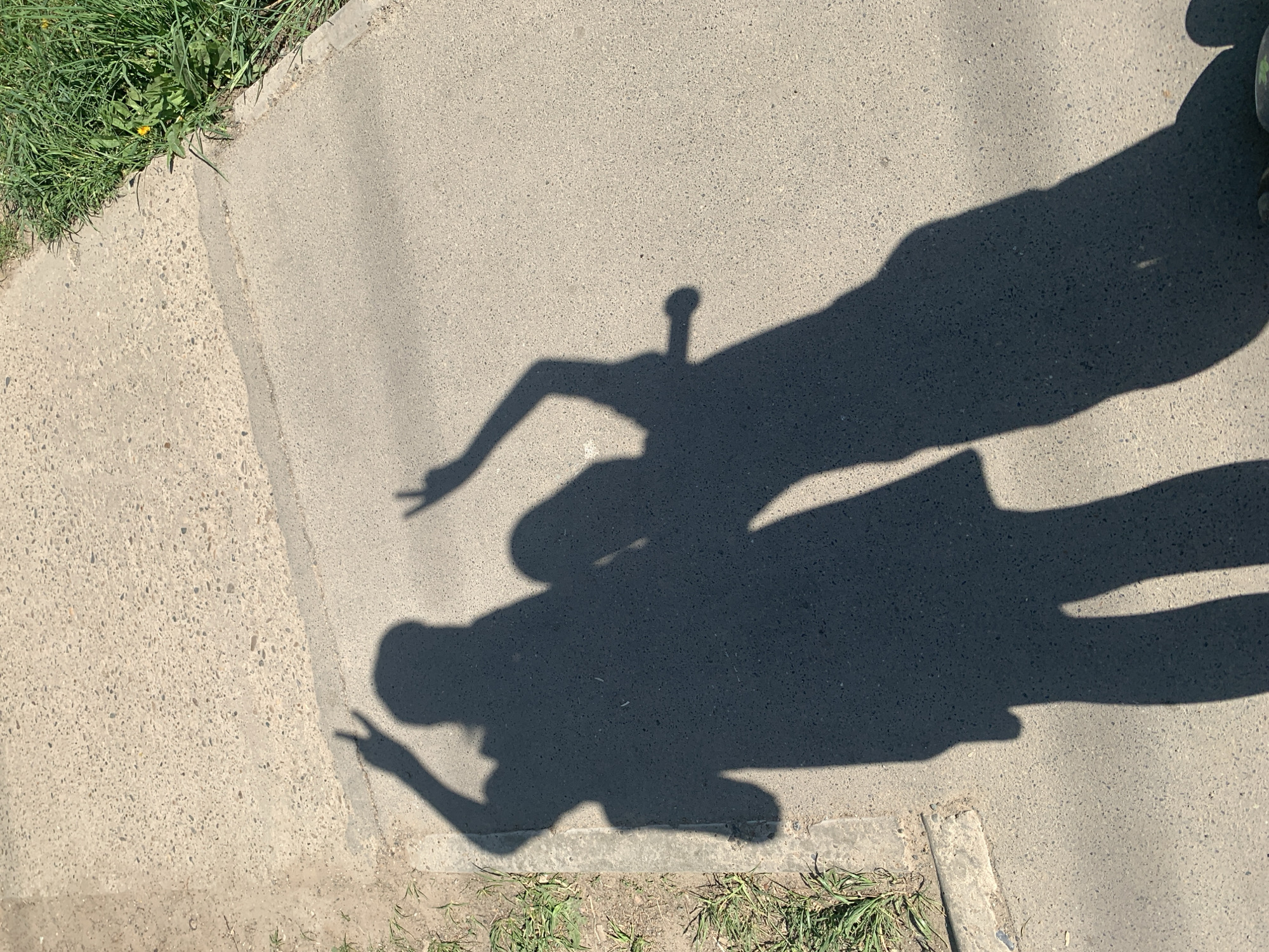 The image shows a segment of a concrete sidewalk in Chaikovsky, Russia, with grass and weeds growing along the edges. The shadows of two people are cast onto the sidewalk. One shadow has an arm raised with fingers in a "peace" sign, while the other shadow's arm is also raised. A small shadow suggests someone is holding an object.
