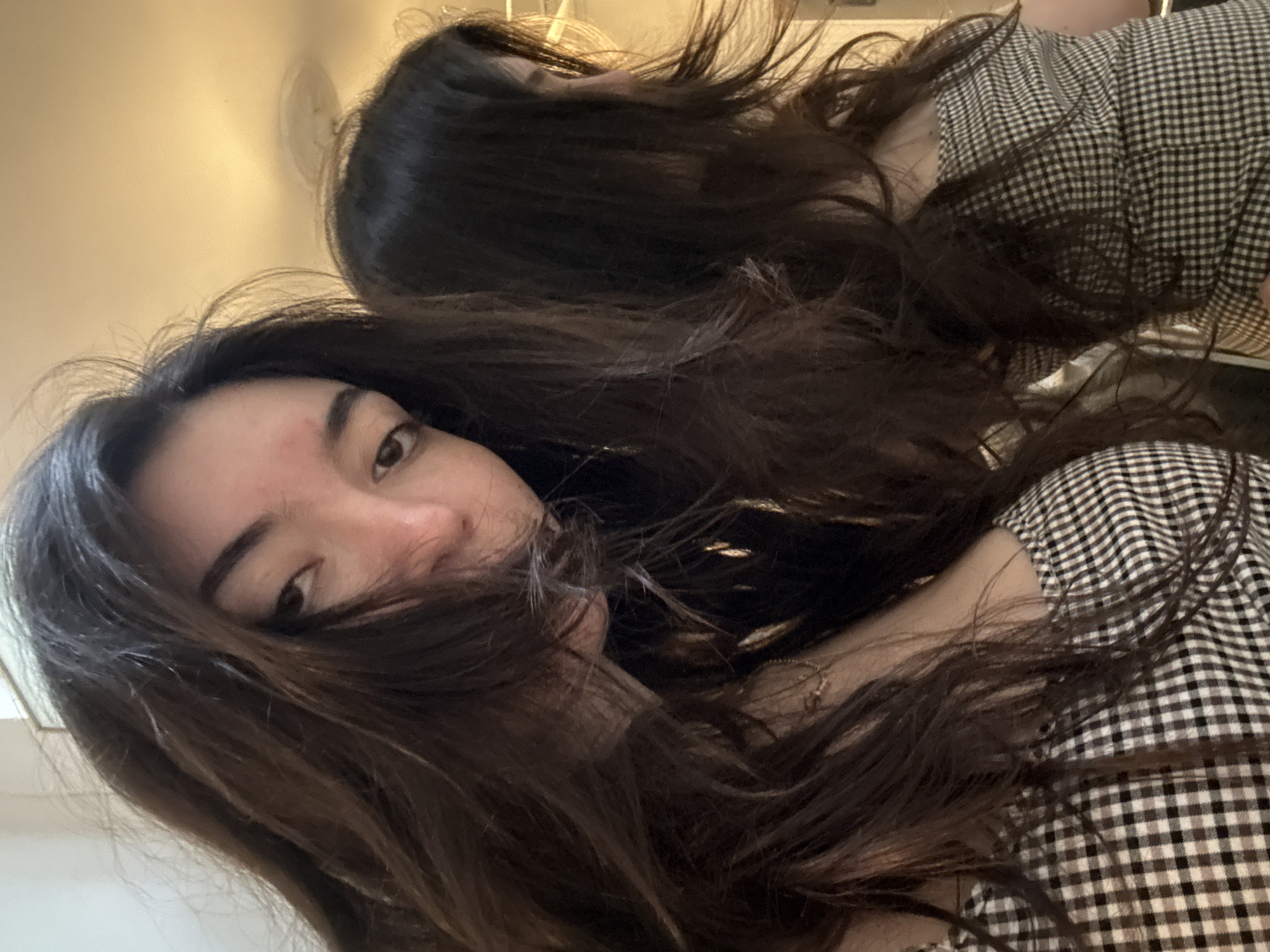 The image shows a young woman with long, dark brown hair. Some strands of her hair cover part of her face. She is wearing a black and white checkered top. A second, blurred image of the woman is in the background. The background is out of focus but appears to be a plain, light-colored wall. The location cannot be determined from the image, but it is not evident that the scene is in New York City.