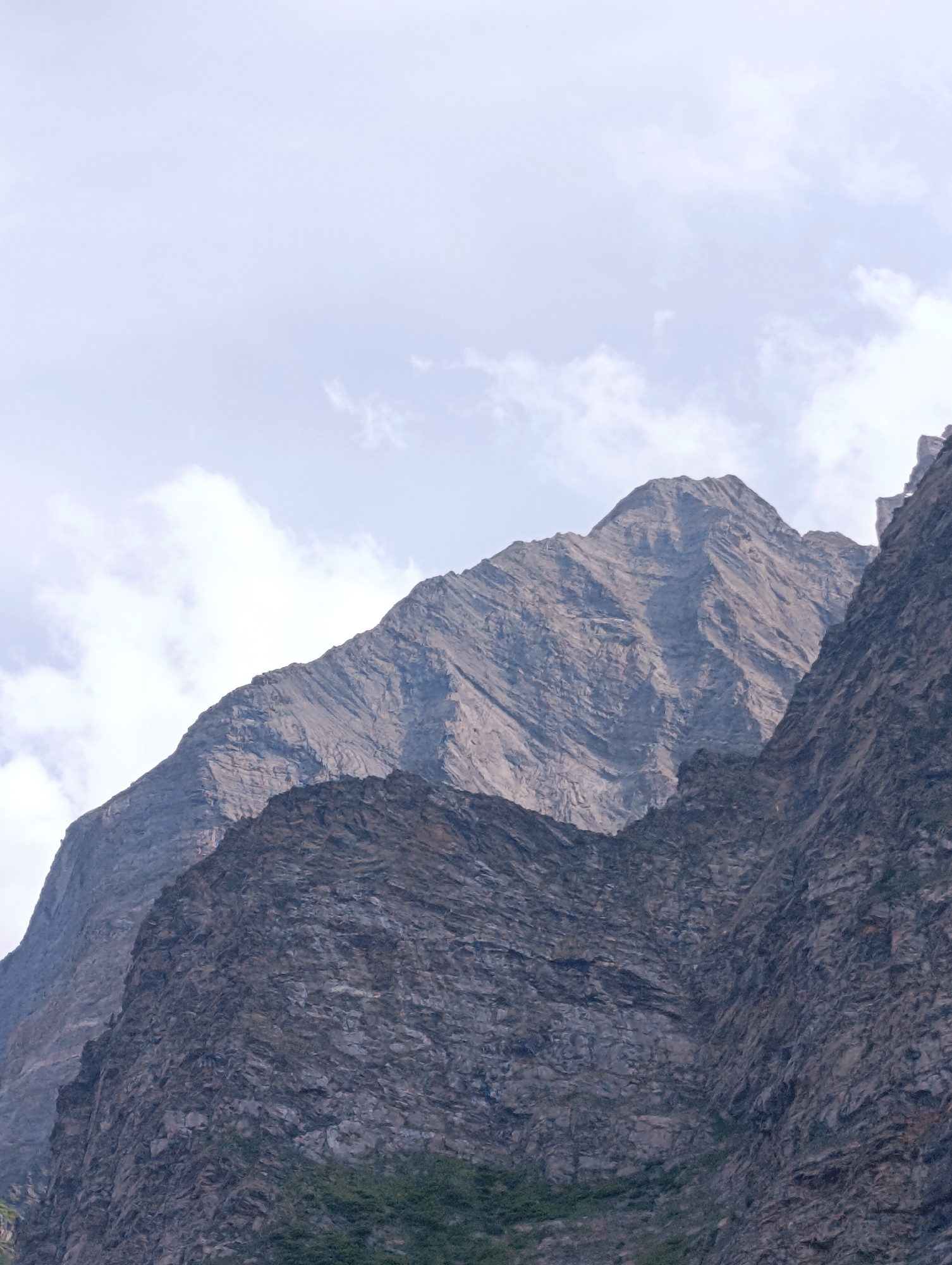 The image shows a close-up view of rugged, rocky mountains under a pale, cloudy sky. The mountains are large and imposing, with steep slopes and visible strata indicating geological layers. The texture of the rock is rough and uneven, with variations in color ranging from dark gray to lighter, weathered tones. In the foreground, a darker, more densely textured rock face is visible, partially obscuring the larger mountain behind it. Sparse green vegetation can be seen at the very bottom of the frame, clinging to the base of the rock. The sky above is a light blue-gray, with soft, scattered clouds. The scene appears to be a natural landscape, likely in a mountainous region, consistent with the description of Tandi, India, which is known for its mountainous terrain. There are no people or man-made objects visible in the image.