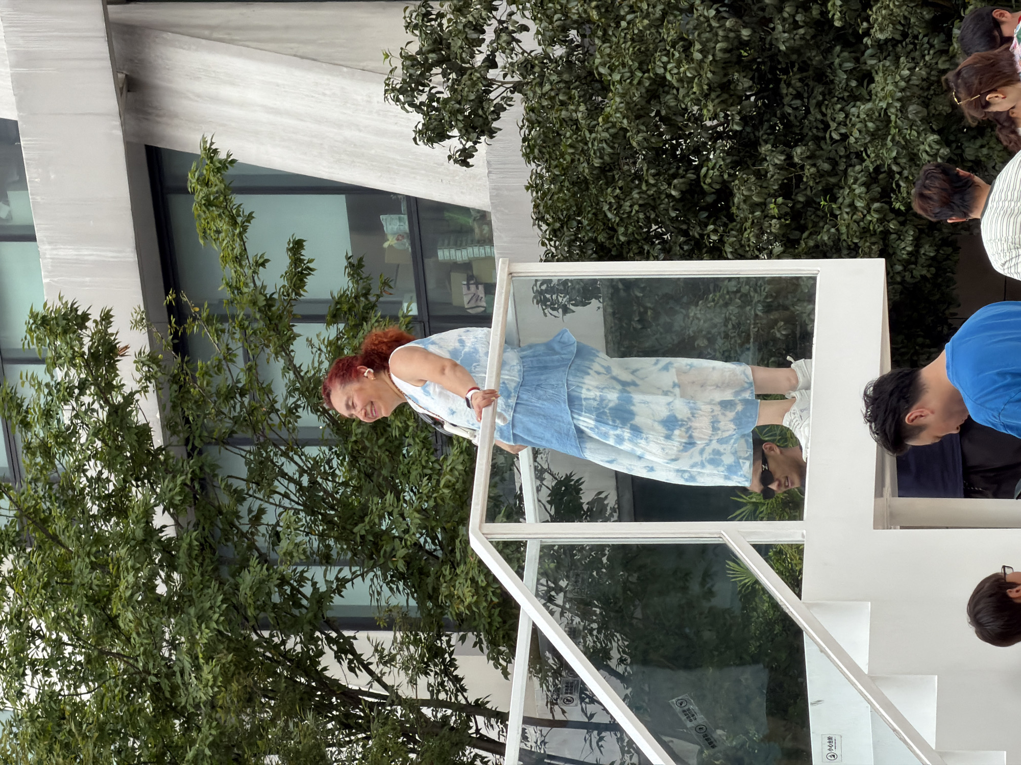 In the Yuzhong District of China, a woman with vibrant red hair is standing on a white multi-level structure outdoors. She is wearing a tie-dye dress in shades of blue and white, a sleeveless top, and white sneakers. The woman is smiling and looking to her left. Behind her, a modern building with large windows and concrete accents is visible, surrounded by lush green trees. Below her, a man wearing sunglasses is looking up at her. In the foreground, other individuals are partially visible, suggesting a public gathering or event. The environment appears to be an urban outdoor space with a contemporary architectural design.