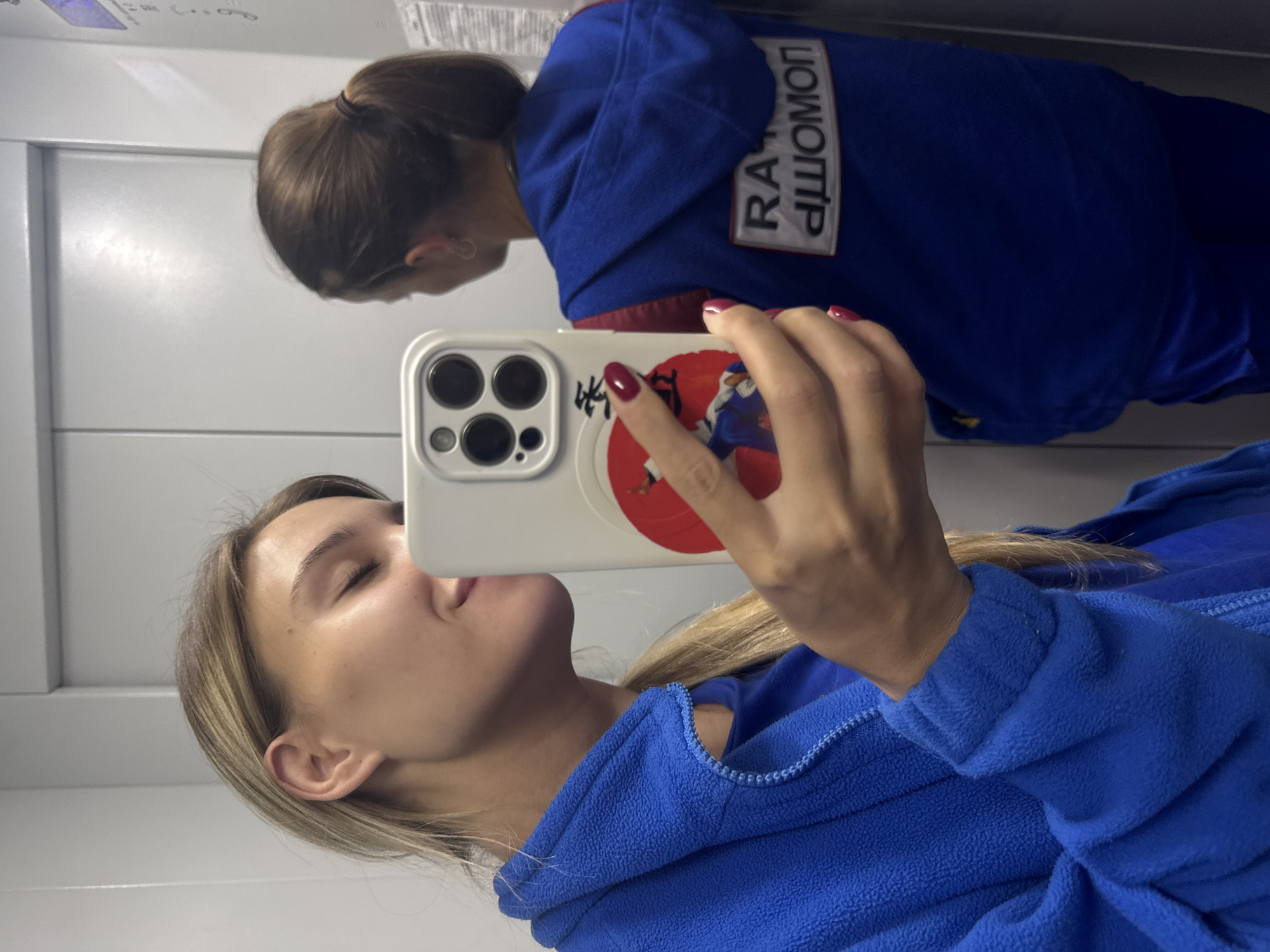 In an elevator in Chaikovsky, Russia, two young women stand facing a mirrored wall, taking a selfie. The woman in the foreground, on the left, is captured from the chest up, holding her phone to take a picture of herself and the woman behind her. She has blonde hair pulled back in a ponytail, is wearing a blue fleece jacket zipped up, and is looking at her phone screen with her eyes closed. The woman behind her, on the right, is seen from the back, her dark brown hair tied in a ponytail. She is wearing a blue hoodie or vest over a red garment. The elevator walls are a neutral grey, and a panel with some writing is visible on the right wall.