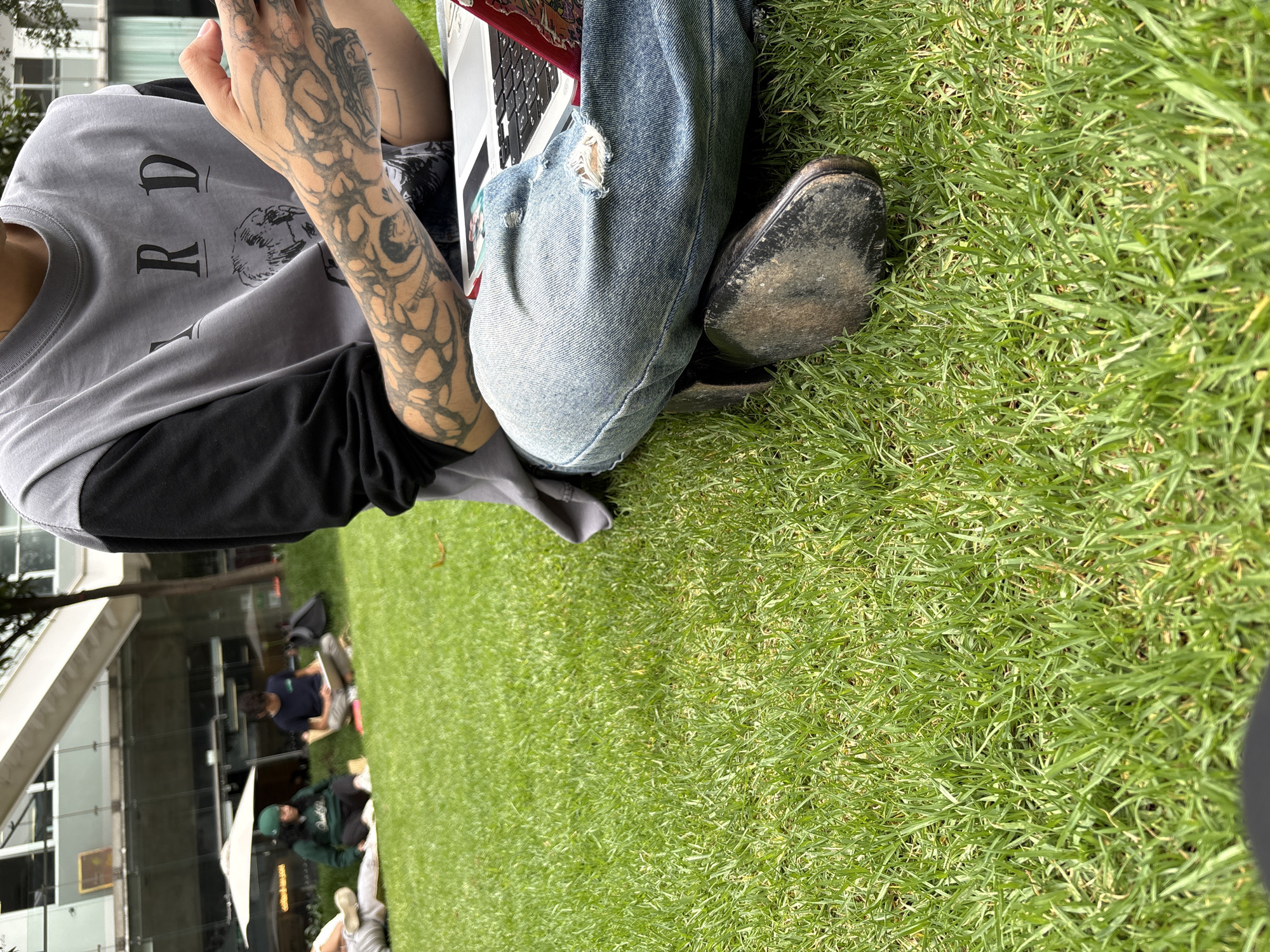A medium eye-level shot reveals a person seated on a grassy area with other individuals scattered in the background, partially obscured by a building. The subject in the foreground is wearing a grey, long-sleeved shirt over a black shirt, blue jeans with a distressed area near the knee, and black leather boots. Tattoos are visible on the left arm and hand. The person appears to be using a laptop. The environment consists of a well-maintained lawn and a blurred background of a building with glass windows and a group of people sitting or lying on the grass. Based on the style of the building and landscape, this photograph was taken in Mexico City, Mexico.