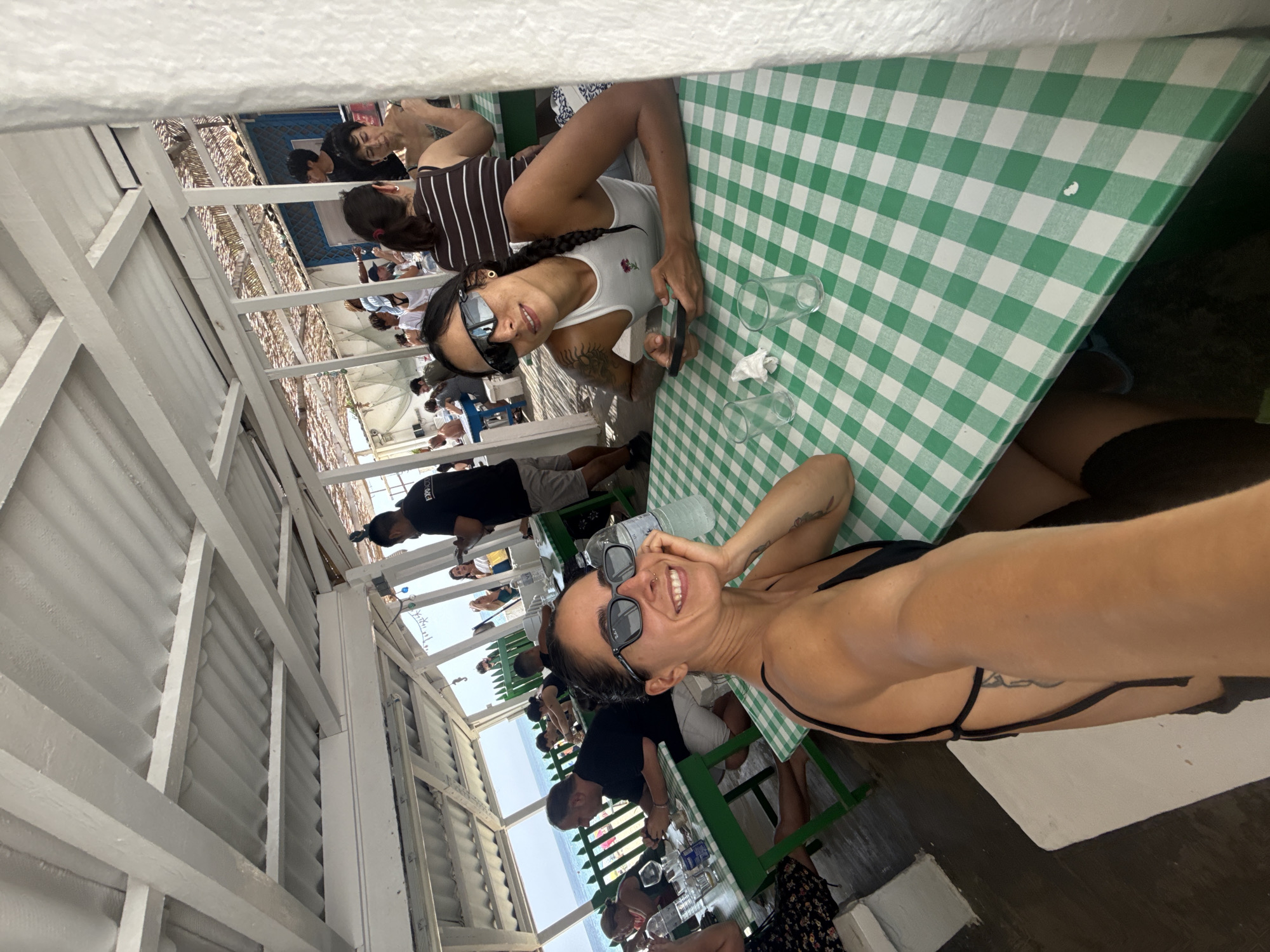 This is a medium shot, eye-level, slightly low-angle, outdoor, full-color photograph of two women dining at a seaside restaurant in Montgat, Spain. The restaurant appears to be open-air, with a white corrugated metal roof supported by white wooden beams. The tables are covered with green and white checkered tablecloths.
In the foreground, on the right, is a woman wearing sunglasses, a white tank top, and a black bikini top. She has a tattoo on her left arm and is holding a smartphone. She is looking at the camera and smiling. To her left, closer to the viewer, is another woman, also wearing sunglasses and a black bikini top. She is also looking at the camera and smiling.
In the background, other diners are visible at various tables. A waiter in a black shirt and shorts is standing near the back of the restaurant. Beyond the restaurant, a green fence separates it from the beach, and the Mediterranean Sea is visible, with a clear blue sky above. The overall atmosphere is casual and relaxed, suggesting a pleasant dining experience by the sea.