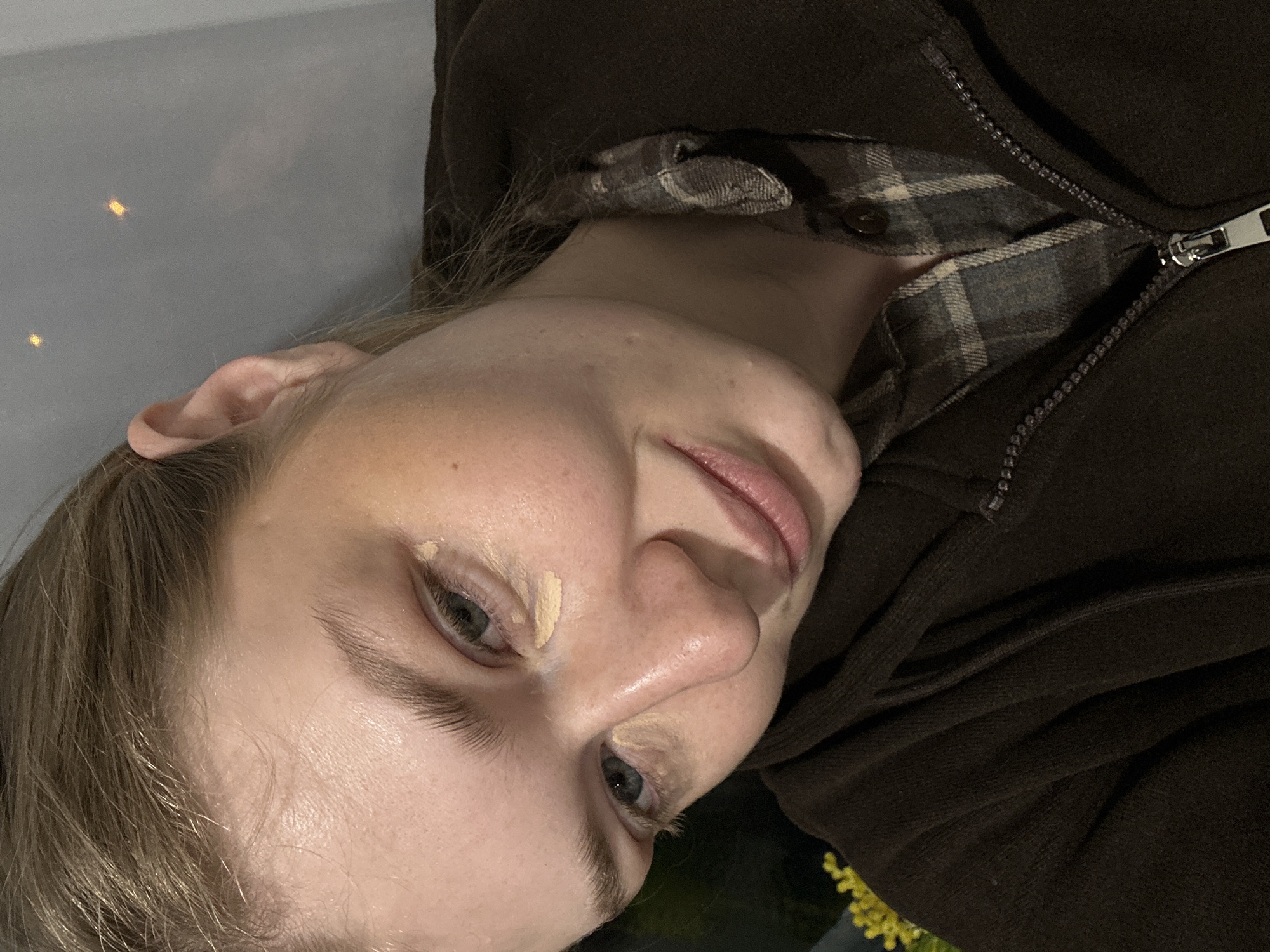 This image presents a close-up, head-and-shoulders portrait of a young adult individual, appearing female, located in Kazan, Russia. The individual has light skin, light brown hair styled away from the face, and blue-grey eyes. The subject is wearing a dark brown, possibly hooded, zip-up garment with a partially visible silver zipper, over a grey and beige plaid shirt. A light-toned, yellowish concealer product is distinctly visible in two applications beneath the subject's left eye (viewer's right), one at the inner corner and another along the lower lash line. A larger, more blended application of the same product is present on the upper right cheekbone area (viewer's left). The subject displays a subtle, closed-mouth smile. The background is dark and out of focus, suggesting an indoor setting. Blurred yellow light sources are visible on the far left, and two small, bright, rectangular reflections are present on a smooth, grey surface behind the subject's right ear. The perspective is slightly elevated, looking down at the subject, whose head is tilted.