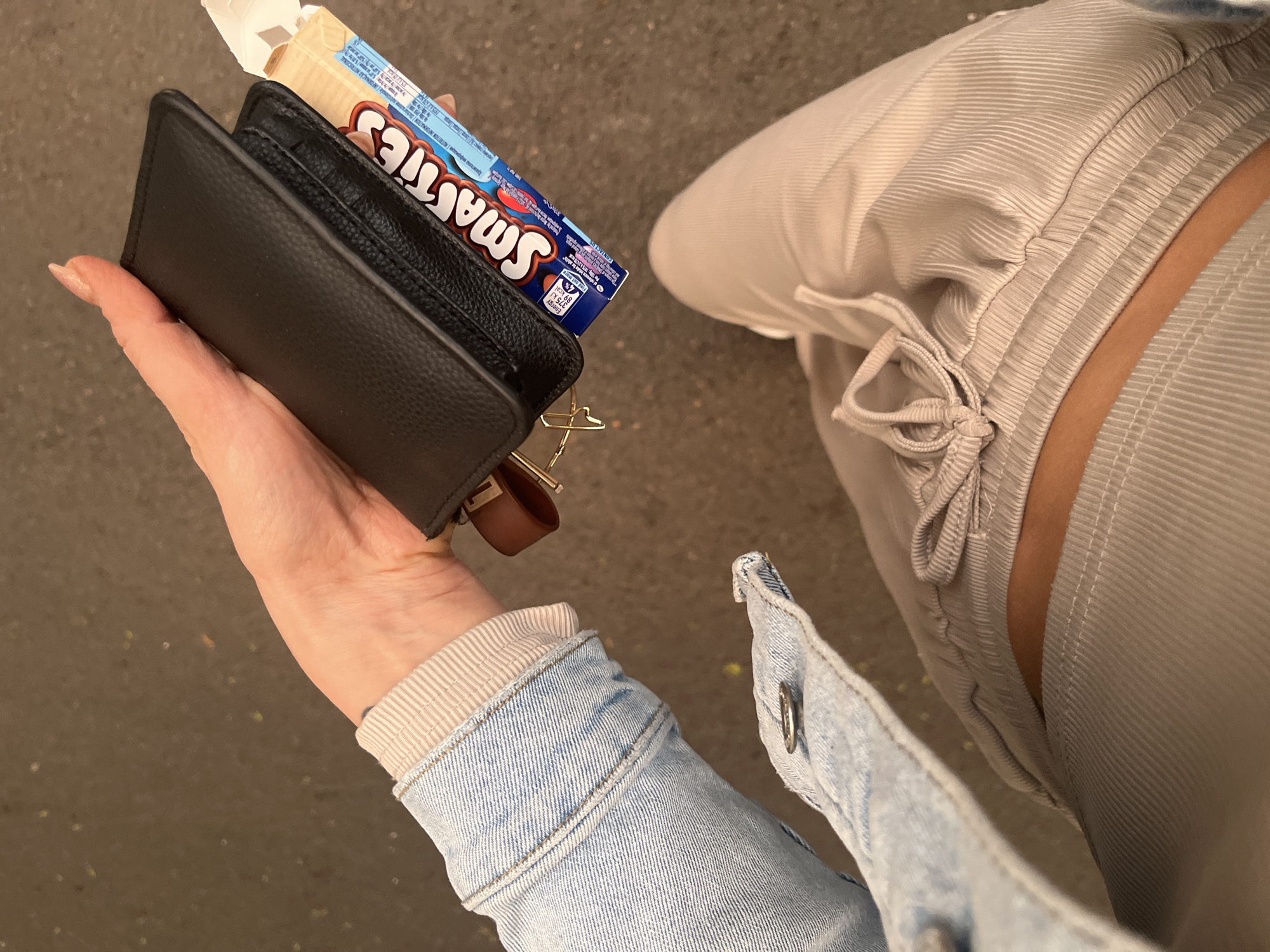 Here is a detailed description of the image:
The image is a close-up, top-down shot that appears to have been taken outdoors. The focus is on a person's hand holding a black leather wallet and a box of Smarties candy. The partially opened candy box reveals the colorful Smarties inside. Attached to the wallet are gold-colored keys and a brown leather keychain.
The person is wearing a light-colored, ribbed pair of pants and a light blue denim jacket with a beige-colored cuff. The ground appears to be asphalt or concrete.
There is no specific information in the image that would identify the location as Varna, Bulgaria.