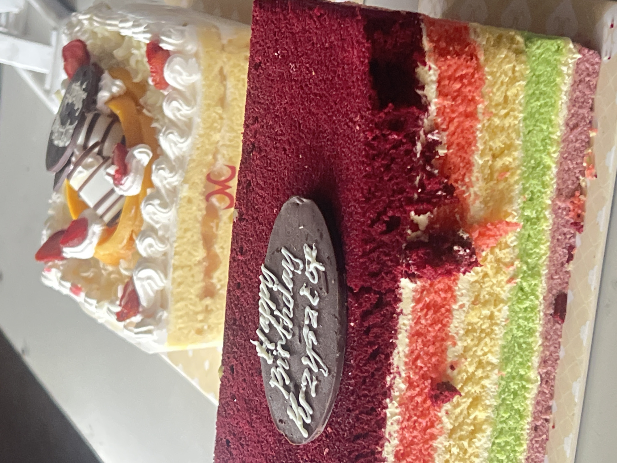 The image shows two cakes. In the foreground is a rectangular cake with layers of red, pink, yellow, and green sponge cake, topped with a dark red crumb coating. An oval chocolate plaque with white icing decoration reads "Happy Birthday Krzysztof". Behind it, to the right, is a square cake with white frosting, decorated with strawberries, peach slices, and chocolate pieces. The cakes appear to be on a white surface, possibly a table. The background is out of focus and shows hints of a white structure. There is no indication of the city of Denpasar, Indonesia, or any specific location.