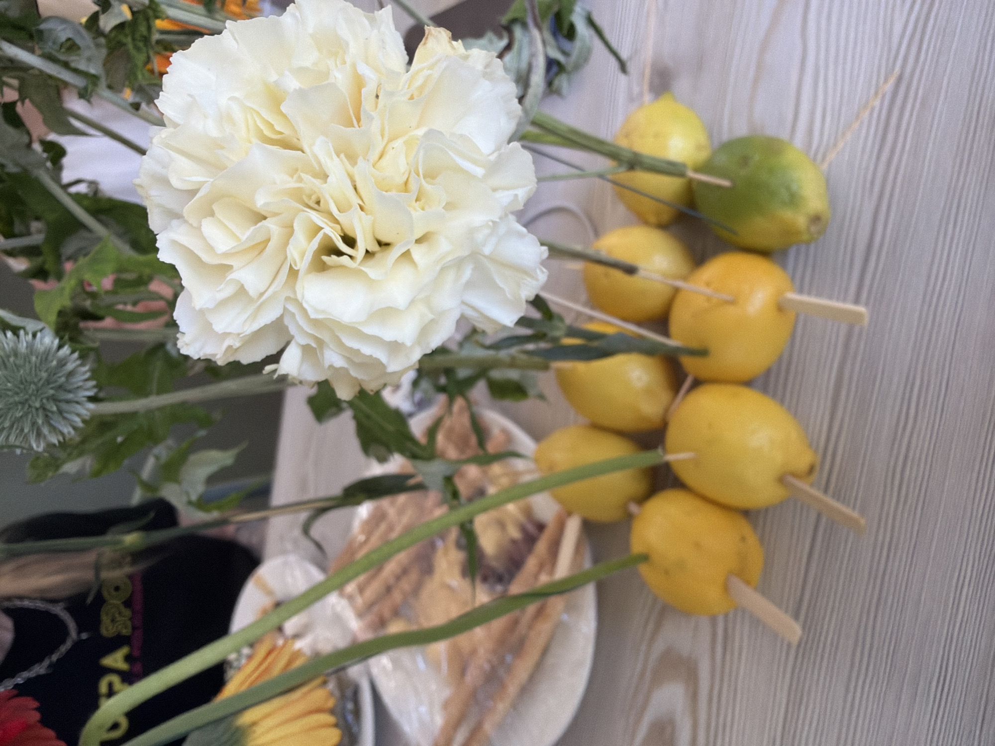 A close-up, eye-level shot captures a table arrangement of decorative elements. In the foreground, several bright yellow lemons, impaled with light-colored wooden skewers, are arranged in a cluster. One lemon on the right is a muted green. These lemons appear to be part of a larger floral display, as several thin green stems rise from behind and between them, supporting a large, creamy white carnation at the center of the composition. To the left of the lemons and carnation, a white plate is visible, holding a transparent plastic bag filled with long, thin breadsticks. In the background, slightly out of focus, a person is partially visible wearing a black t-shirt with yellow text and a silver chain necklace. The surface of the table is made of light-colored wood with a horizontal grain. The scene appears to be indoors, likely a domestic setting.