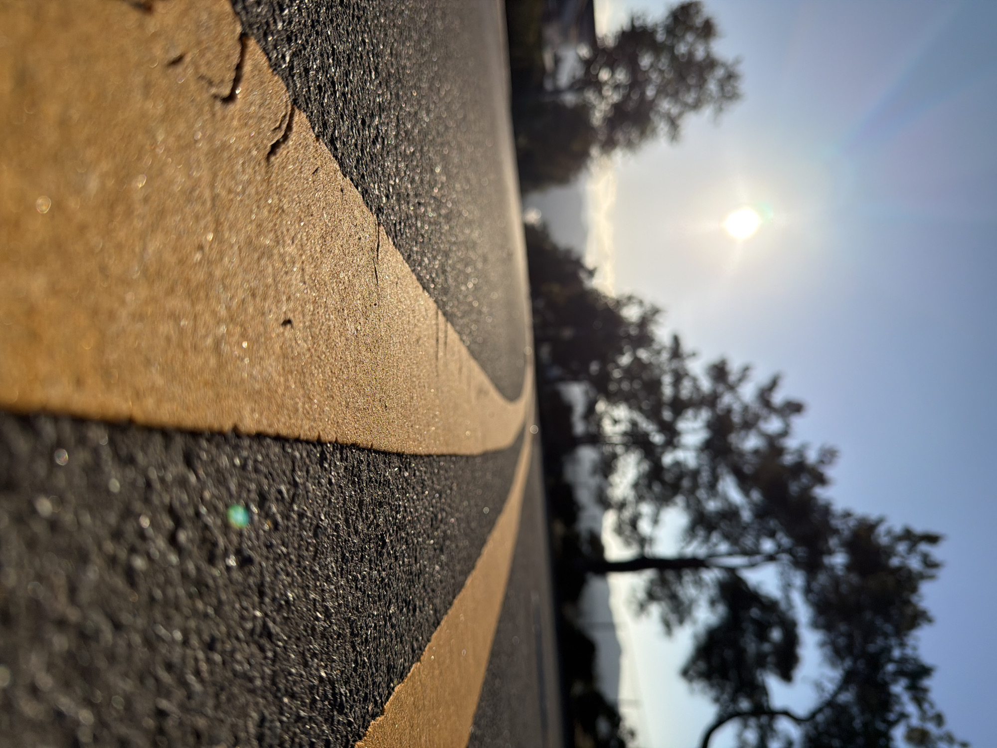 The image shows a close-up, low-angle view of a road with yellow double lines. The road surface is asphalt, and the yellow lines are brightly colored and appear to be slightly textured. The sun is visible in the upper left quadrant of the image, creating a lens flare and casting a warm glow over the scene. In the background, blurred trees and a clear sky suggest an outdoor setting. The scene is likely located on a road in Aliso Viejo, California, United States, given the typical landscape and road markings found in the area. There are no people or animals visible in the image. The focus is on the road and the play of light and shadow on its surface.