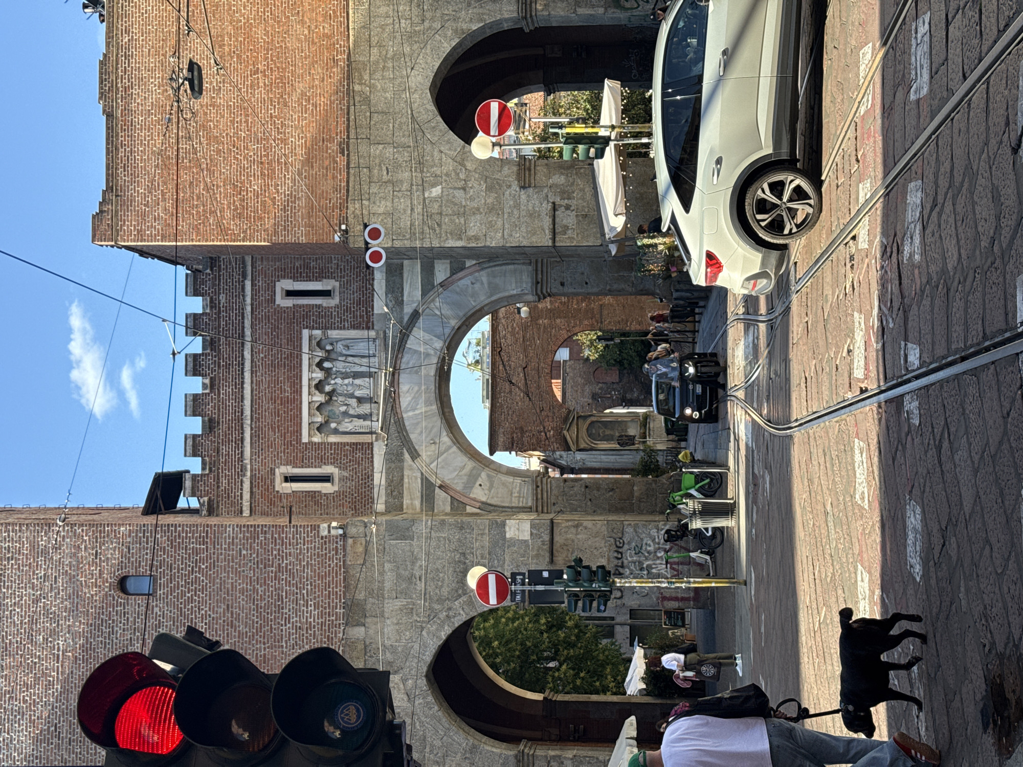 The image depicts a street scene in Milan, Italy, featuring the Porta Ticinese. The gate is constructed of brick and stone and consists of three arches. A white car is parked to the right of the gate. Other vehicles and pedestrians are present. A person is walking a black dog in the foreground. Traffic lights are visible, and one is showing a red light. Tram tracks run along the cobblestone street.