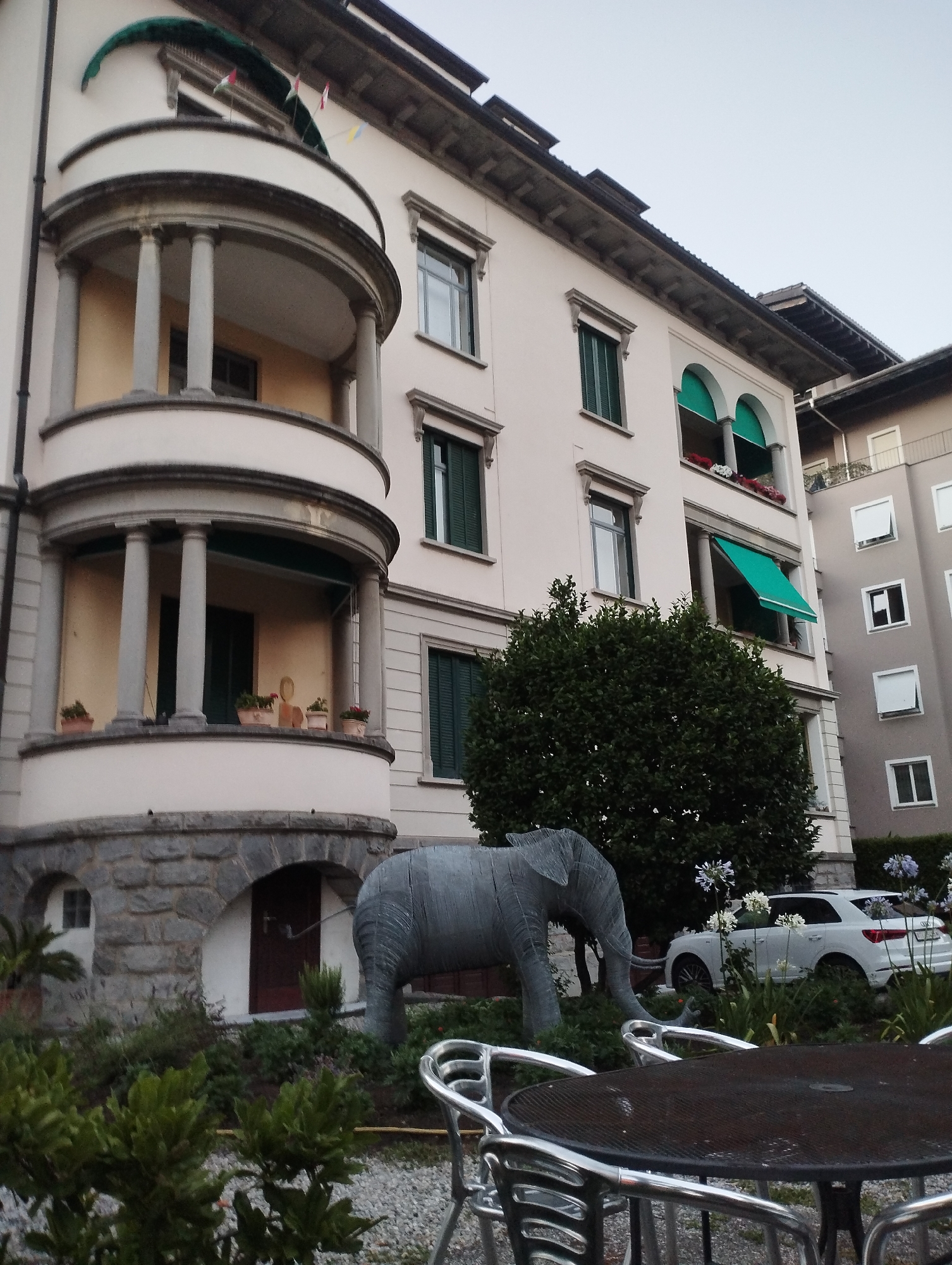 The image shows a view of a building in Locarno, Switzerland, with an outdoor seating area in the foreground. A dark-colored metal table with four chairs surrounds it. To the left, there is a gray sculpture of an elephant. Behind the elephant, a white Audi SUV is parked. The building itself is light-colored, with a rounded balcony supported by columns on the left side. The balcony has plants in pots. The building features several windows with dark green shutters, and some awnings are visible. To the right, another building with multiple windows is partially visible. The ground is covered with gravel, and there are various plants and bushes in the scene. The sky is overcast.
