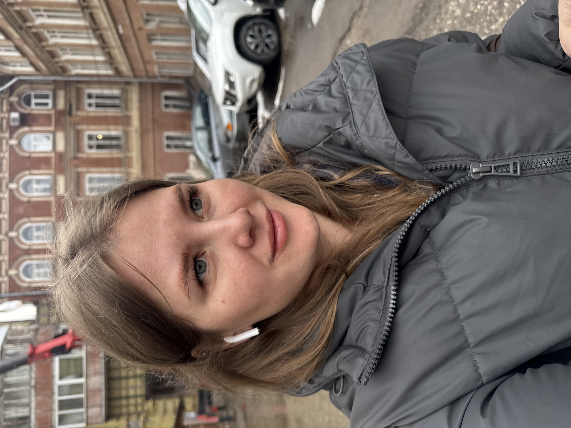 This media file is a portrait-oriented, close-up photograph of a young woman taken outdoors in an urban setting, likely Perm, Russia. The woman is the primary subject, positioned in the foreground, looking directly at the camera with a calm, pleasant expression and a subtle smile. She has light brown, wavy hair that falls around her shoulders and blue-green eyes. A white wireless earbud is visible in her right ear.
She is wearing a dark grey puffer jacket, which is zipped up. The jacket features a prominent zipper pull and a quilted texture on the hood and upper shoulder area.
The background is out of focus, indicating a shallow depth of field, which emphasizes the woman. It reveals an urban street scene. On the left side of the background, there are multi-story buildings constructed from reddish-brown brick, featuring numerous windows with distinct arched tops, suggesting older European architecture. On the right side, several cars are partially visible, including a white SUV with dark wheels and another darker vehicle. A red structure, possibly part of a crane or construction equipment, can be seen in the lower left portion of the background behind her.
The lighting suggests it is daytime, possibly overcast, which provides soft, even illumination on her face. The puffer jacket implies the weather is cool or cold. There is no visible text in the image.