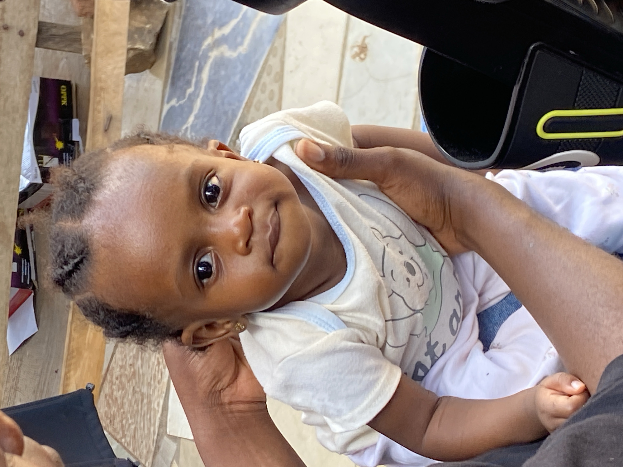 The image features a close-up of a baby being gently held by an adult, likely in Bukuru, Nigeria. The baby, with dark skin and dark, curly hair, is looking directly at the camera with wide, dark eyes and a soft, neutral expression that hints at curiosity. The baby is dressed in a light-colored (cream or off-white) short-sleeved top adorned with a cartoon character, possibly a bear, and some indistinct text below it. Light-colored pants are also visible.
The adult's dark-skinned hands and forearms are seen supporting the baby from below and the sides. Part of the adult's dark-colored clothing is visible in the lower right portion of the frame.
In the upper right, a dark, structured object with bright yellow accent lines is present, possibly part of a stroller, carrier, or another piece of equipment. The background shows a light-colored, patterned tiled or paved surface, suggesting an outdoor setting or a brightly lit indoor space. On the left side of the background, a box is visible with the text "OPPK" clearly printed on it. The scene is well-lit by natural light, indicating it is daytime.